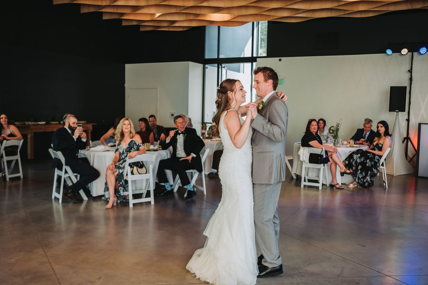 A couple shares their first dance as newlyweds at a rustic indoor wedding reception with guests watching.