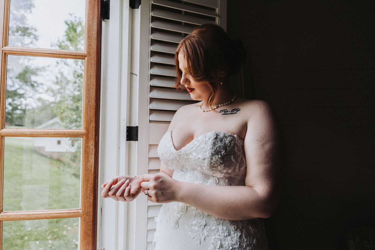A bride in a white lace dress stands thoughtfully by a window in soft natural light.