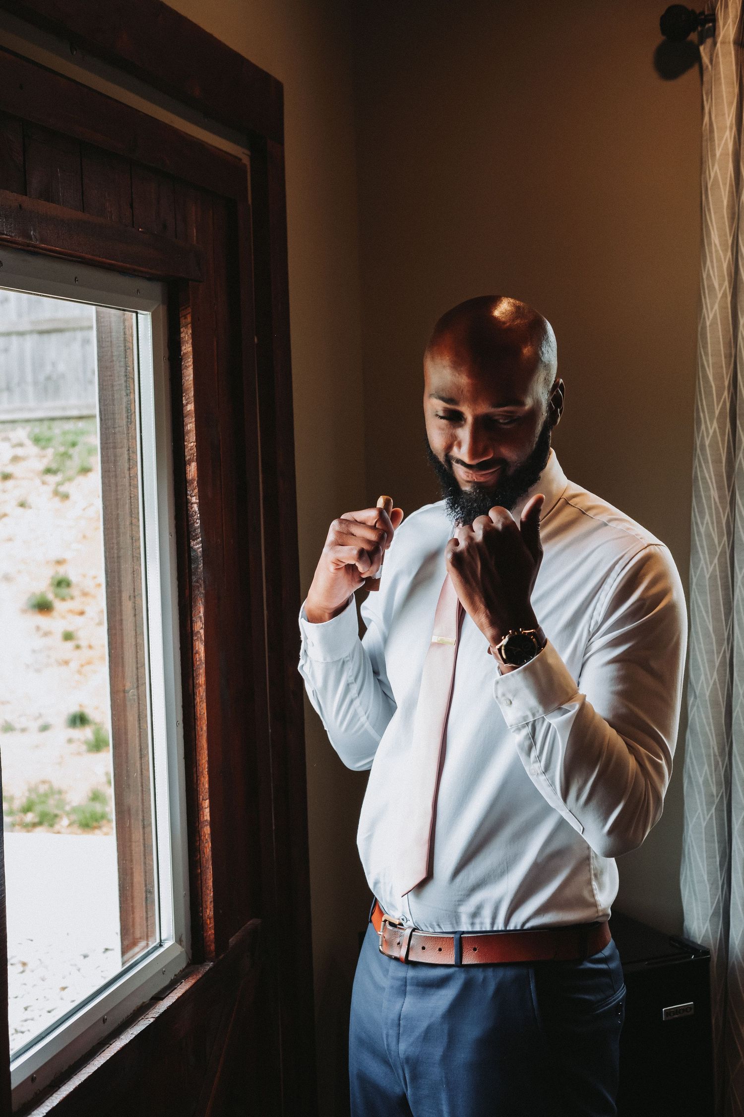 Smiling man in formal attire adjusts his tie while standing near a window in warm lighting.