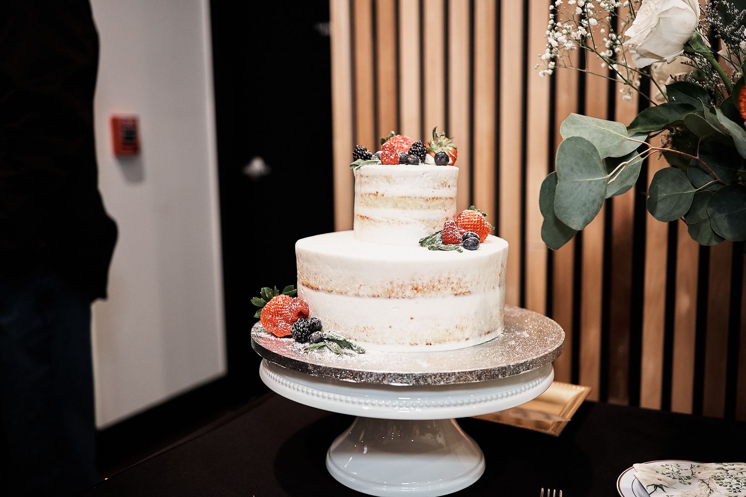 A white three-tiered wedding cake decorated with coral roses and eucalyptus on a pedestal stand against wooden slats.