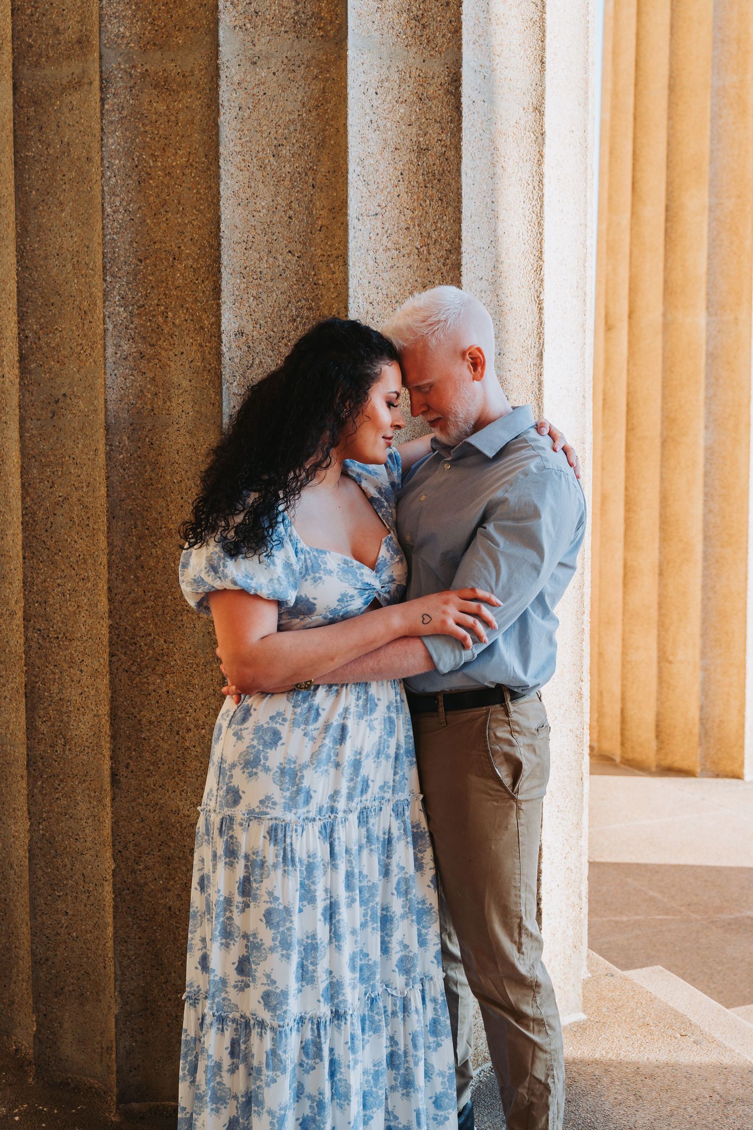 A couple embraces tenderly between stone columns, woman in blue floral dress, man in blue shirt, sharing an intimate moment.