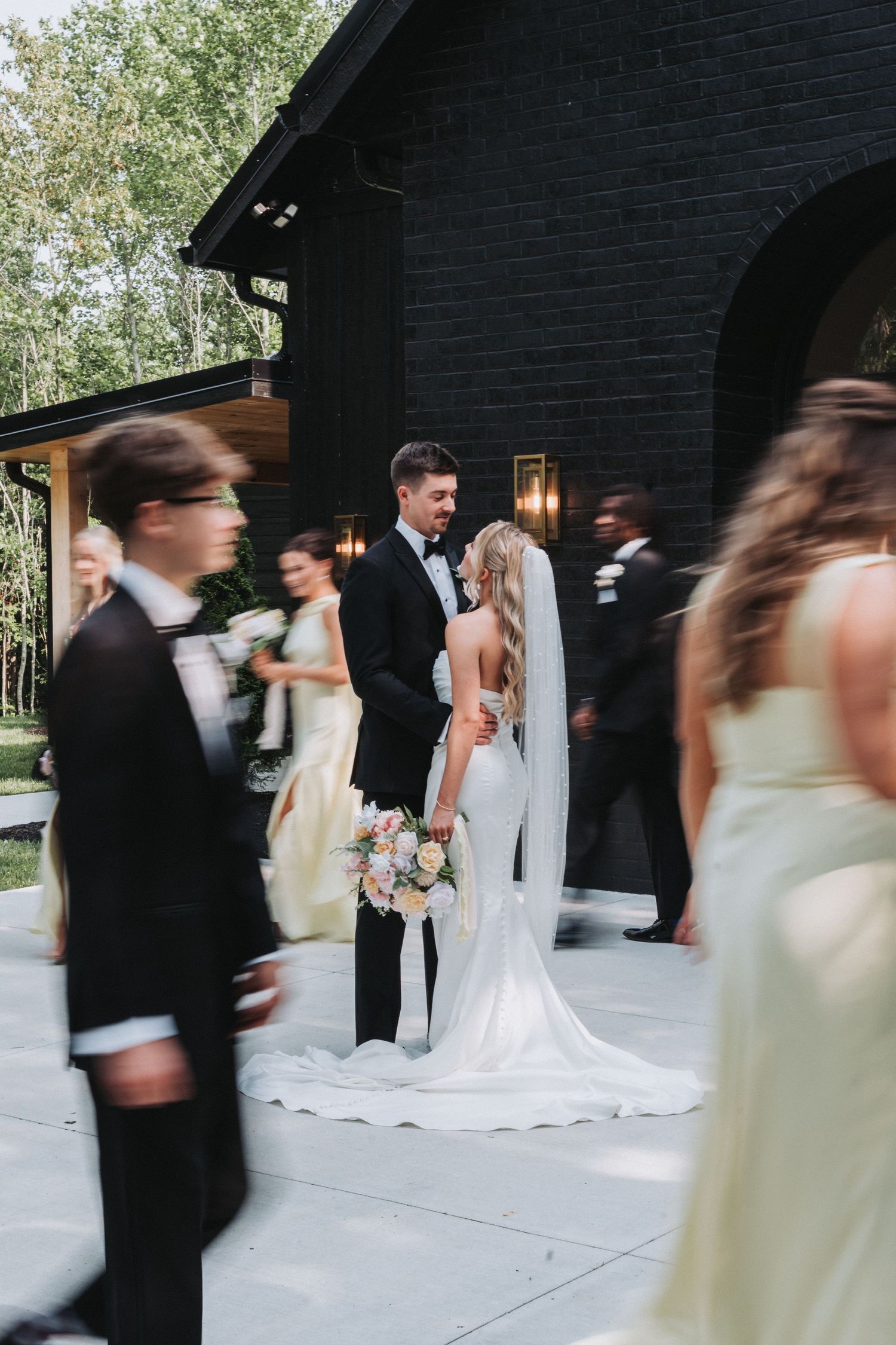Motion blur captures wedding party walking outside black building with tall windows and surrounding greenery.