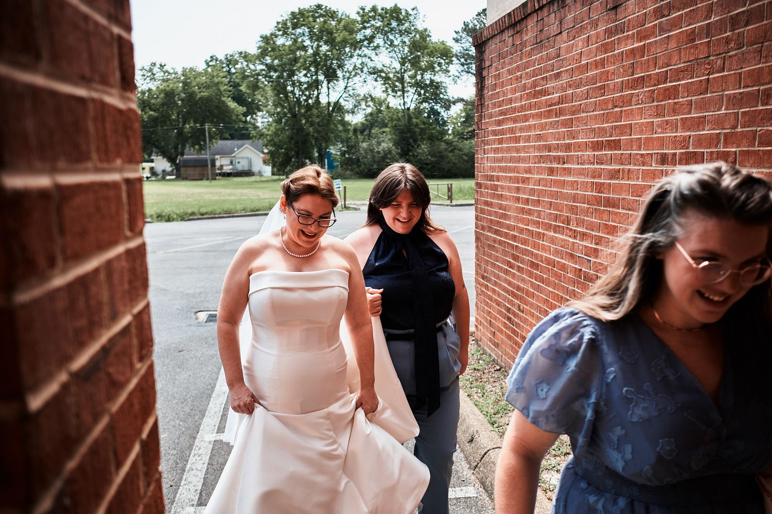 Bride and bridesmaid walk together along a rustic brick building exterior during a wedding celebration.