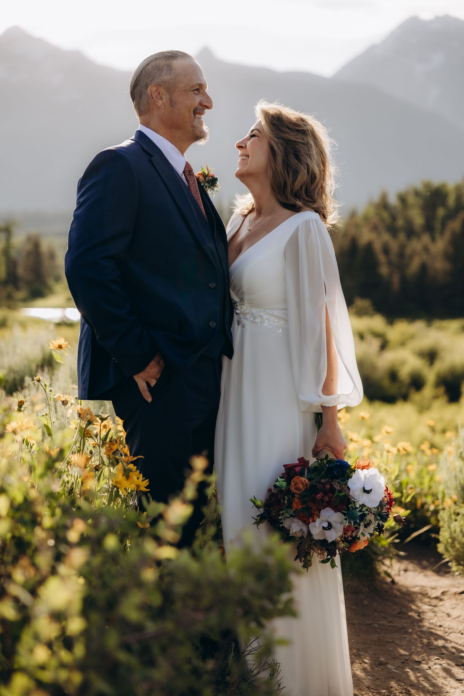 A couple in wedding attire share intimate moments in a sunlit meadow with mountains and wildflowers in the background.