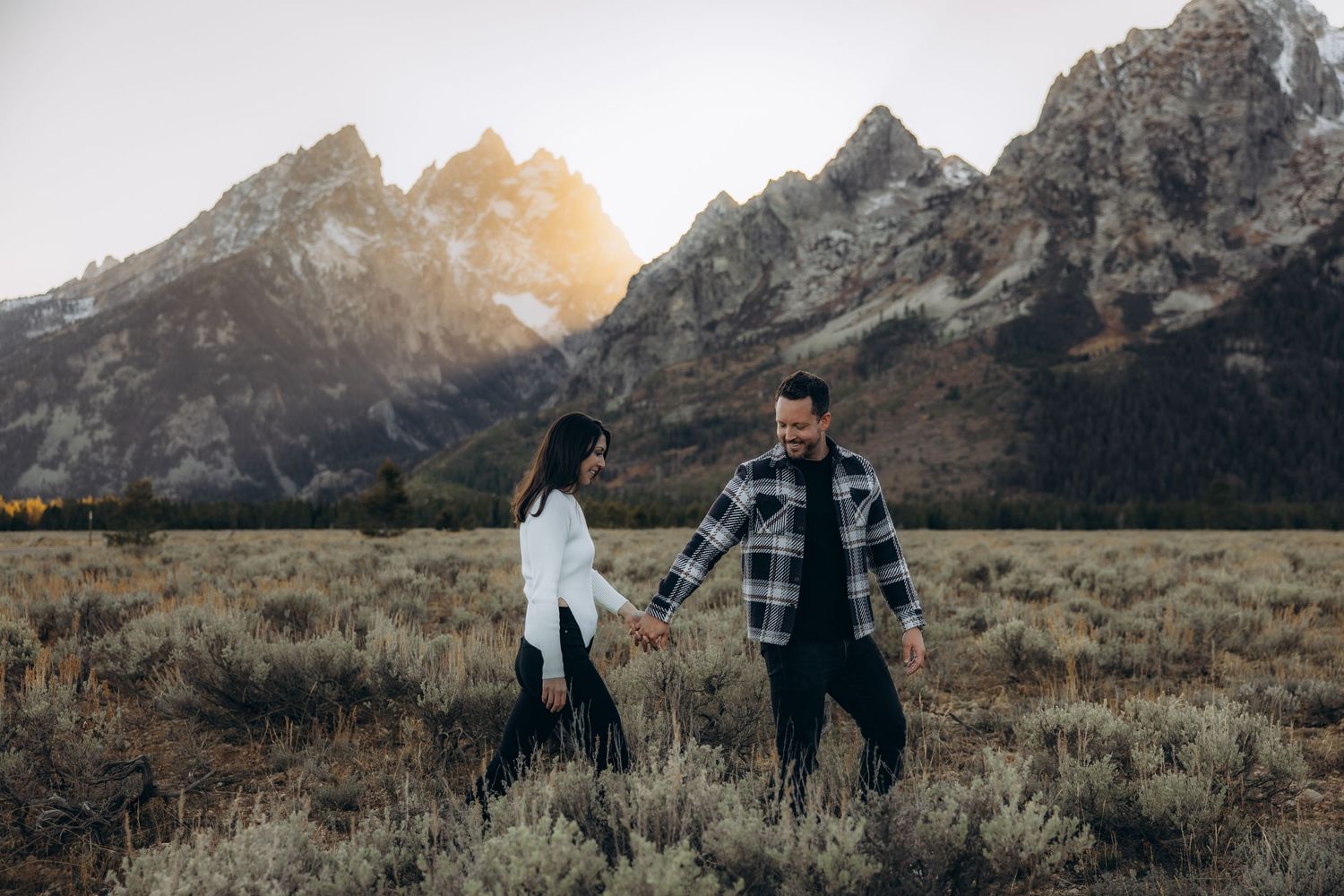 Two people stand in a field of dry grass with dramatic mountain peaks silhouetted against a sunset sky in Grand Teton.