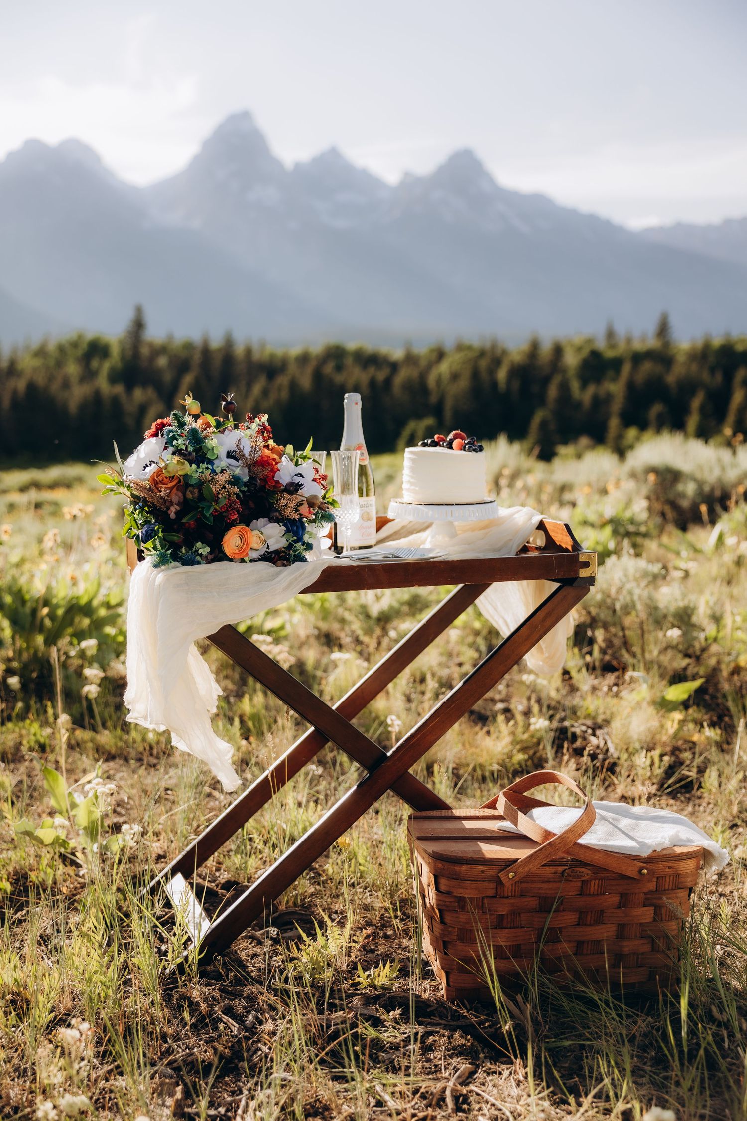 Rustic wooden picnic setup with floral arrangements and white decor against a scenic mountain backdrop in golden meadow.