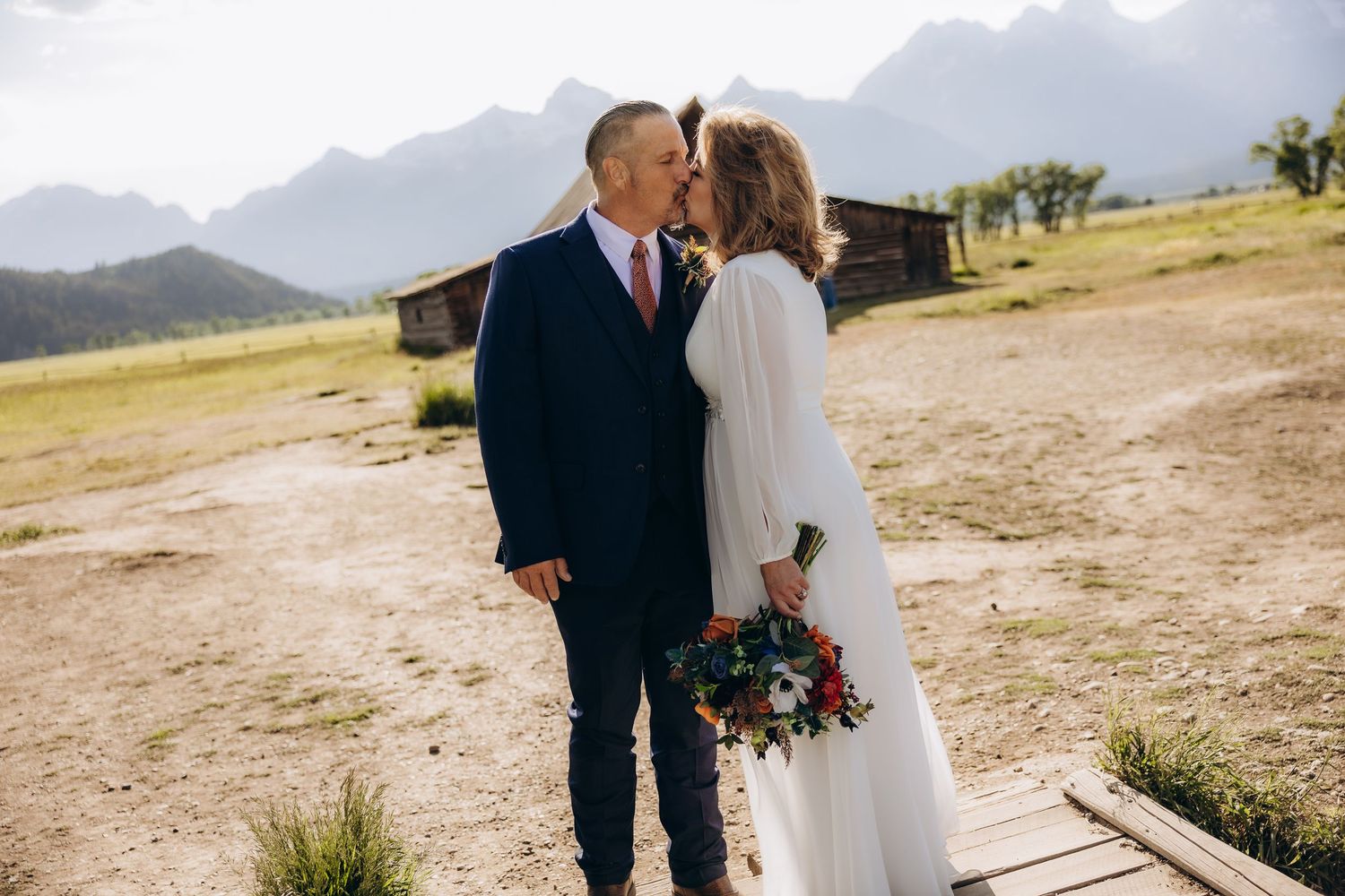 A couple shares a romantic moment on a scenic mountain trail with sunlit meadows and peaks in the background.