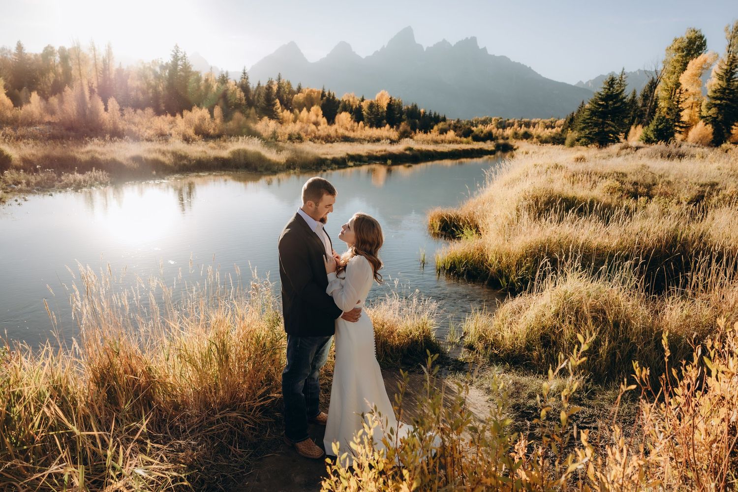 A couple in wedding attire embraces at sunset near a lake with mountains and golden grass in the background.