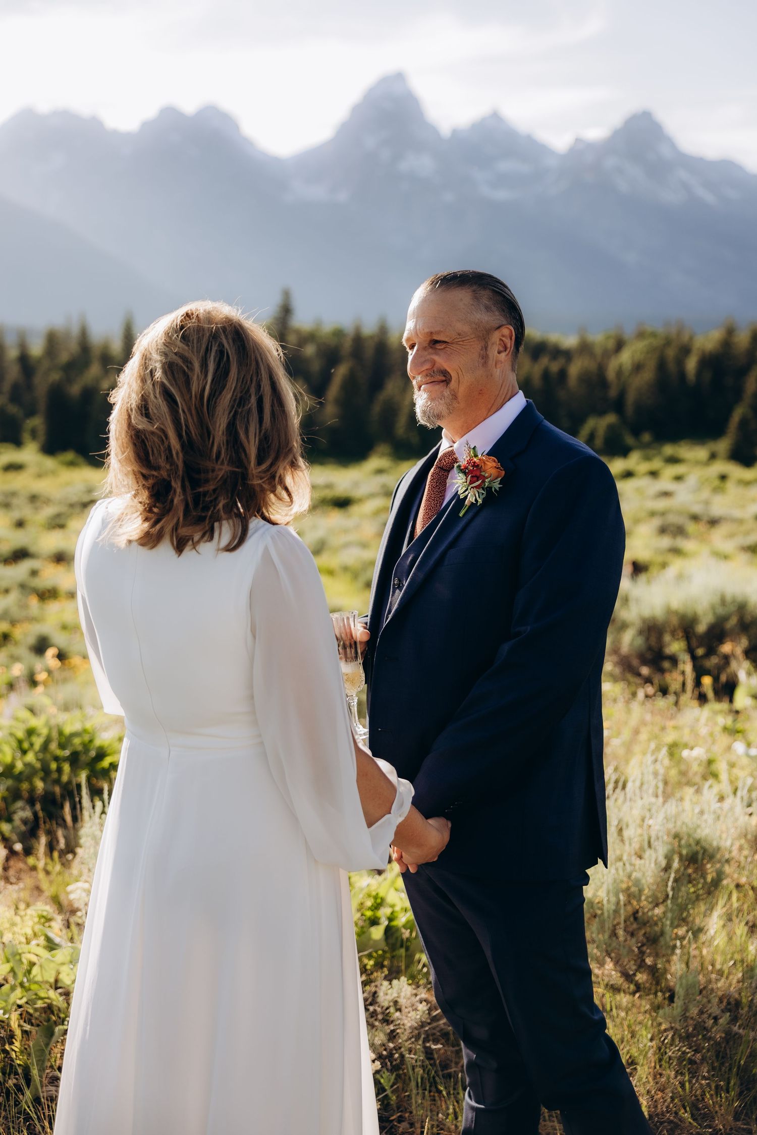 A couple shares an intimate moment in a field with majestic mountain peaks and evergreen forest in the background.