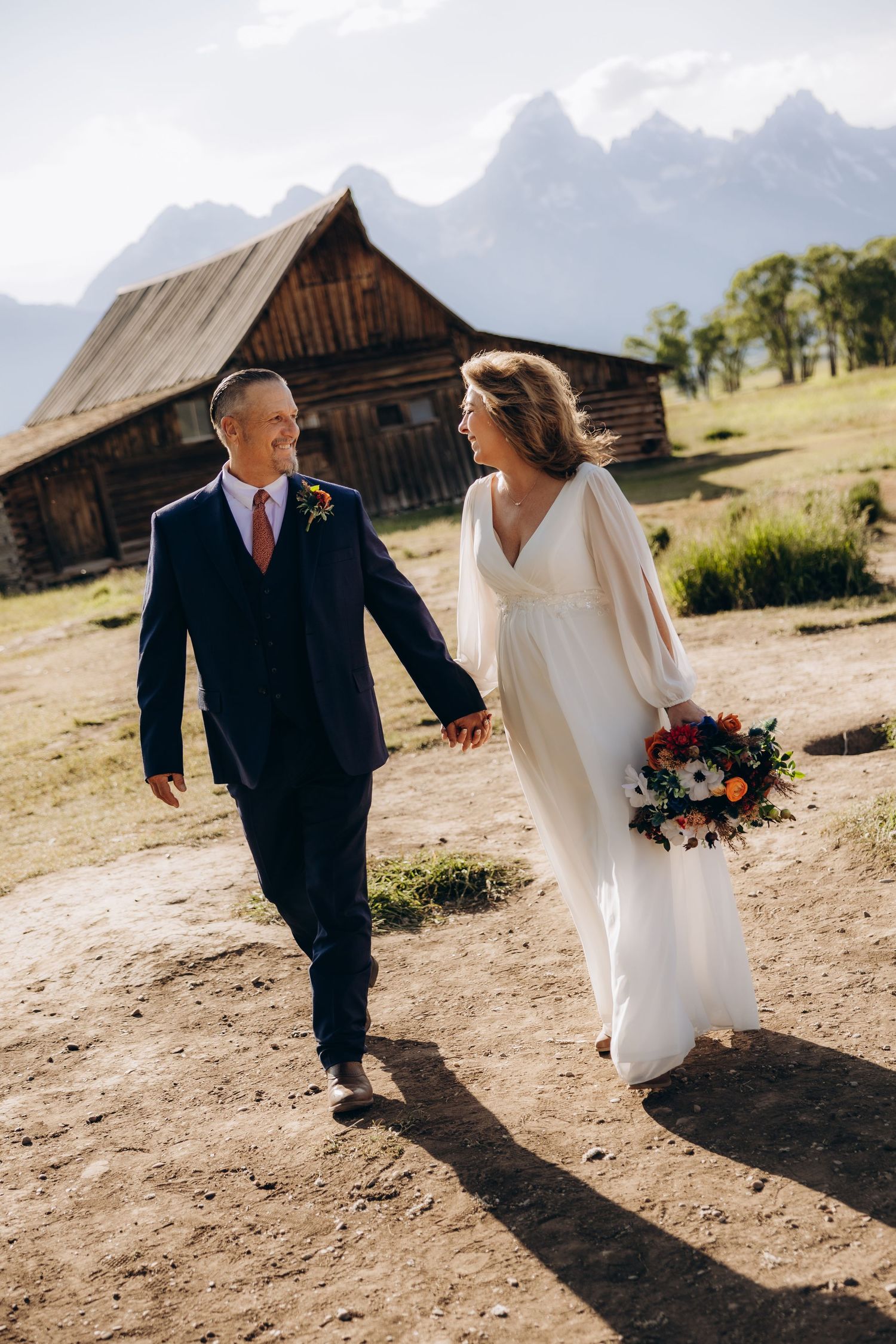 A couple in wedding attire walks hand in hand near a rustic barn with majestic mountains in the background.