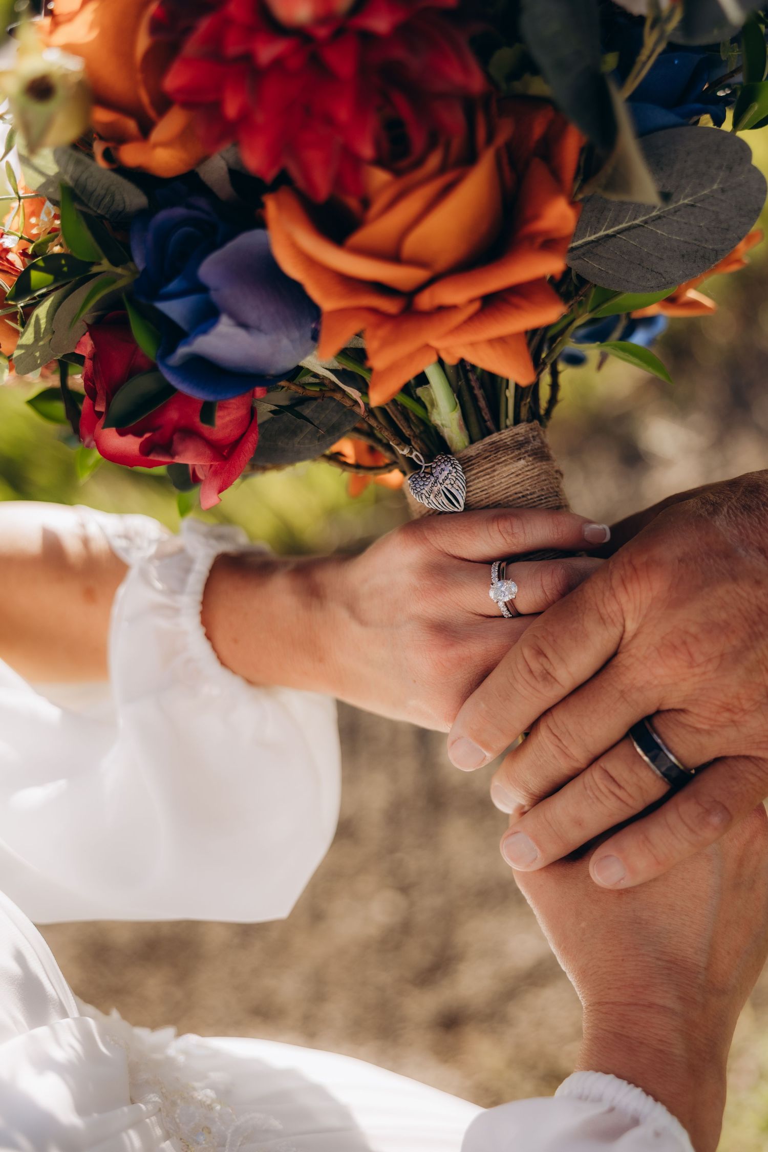 Close-up of hands with wedding rings holding a vibrant autumn-colored bouquet with orange and burgundy flowers.