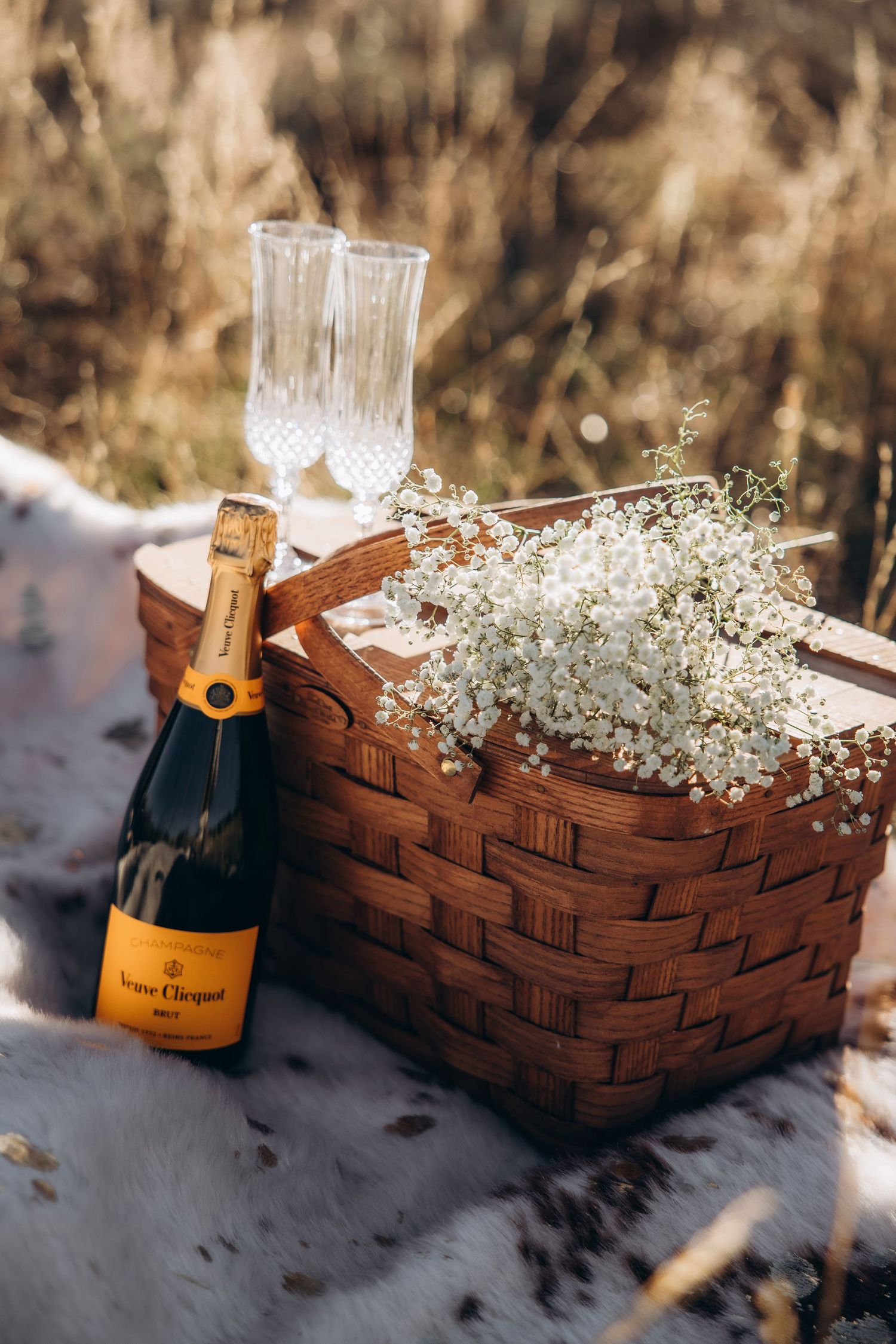 A bottle of Veuve Clicquot champagne with crystal glasses and baby's breath flowers in a woven basket on a snowy surface.