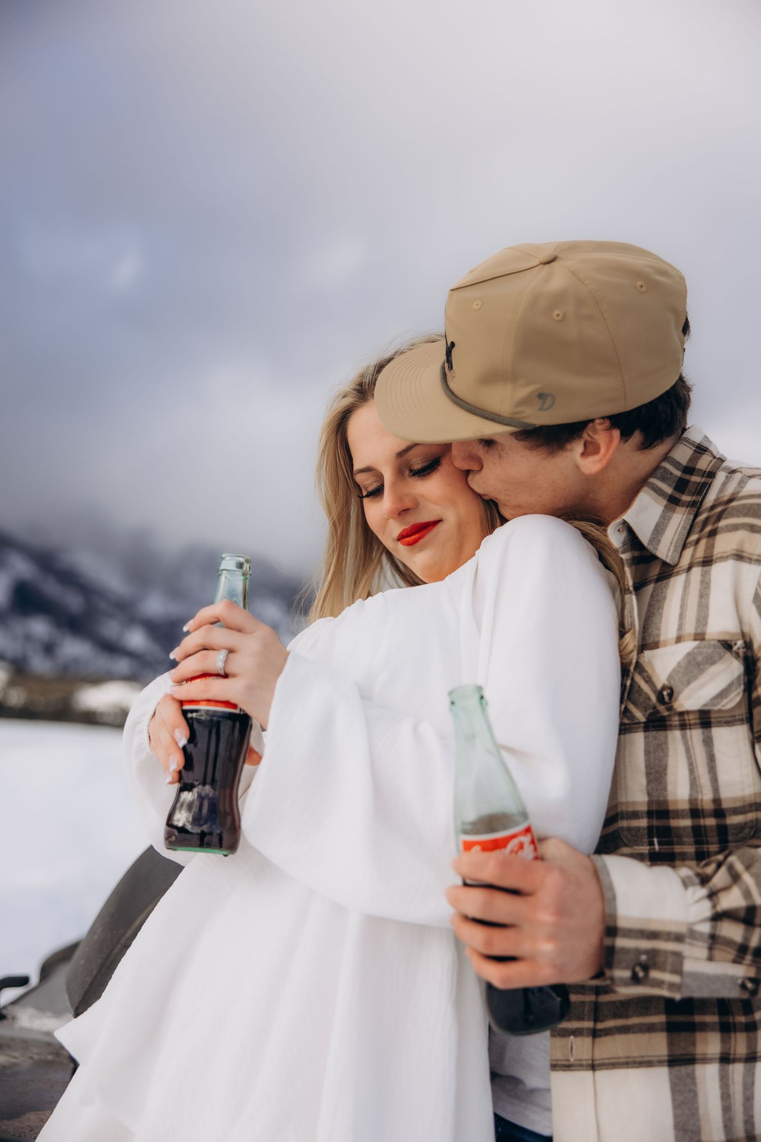 A couple shares an intimate moment in the snow while holding glass bottles against a mountainous winter backdrop.