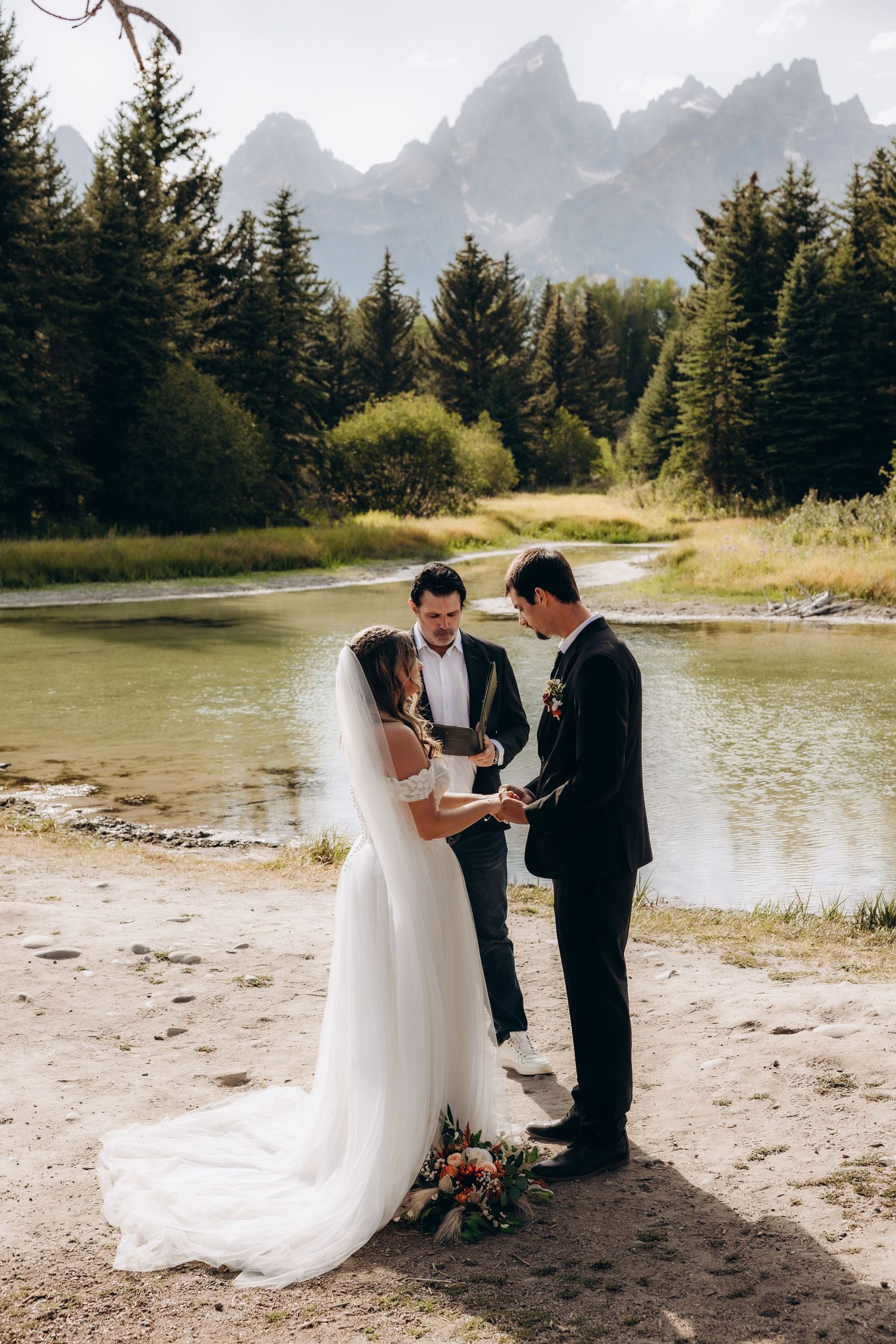 A mountain wedding ceremony takes place on a serene beach surrounded by evergreen trees and dramatic peaks.
