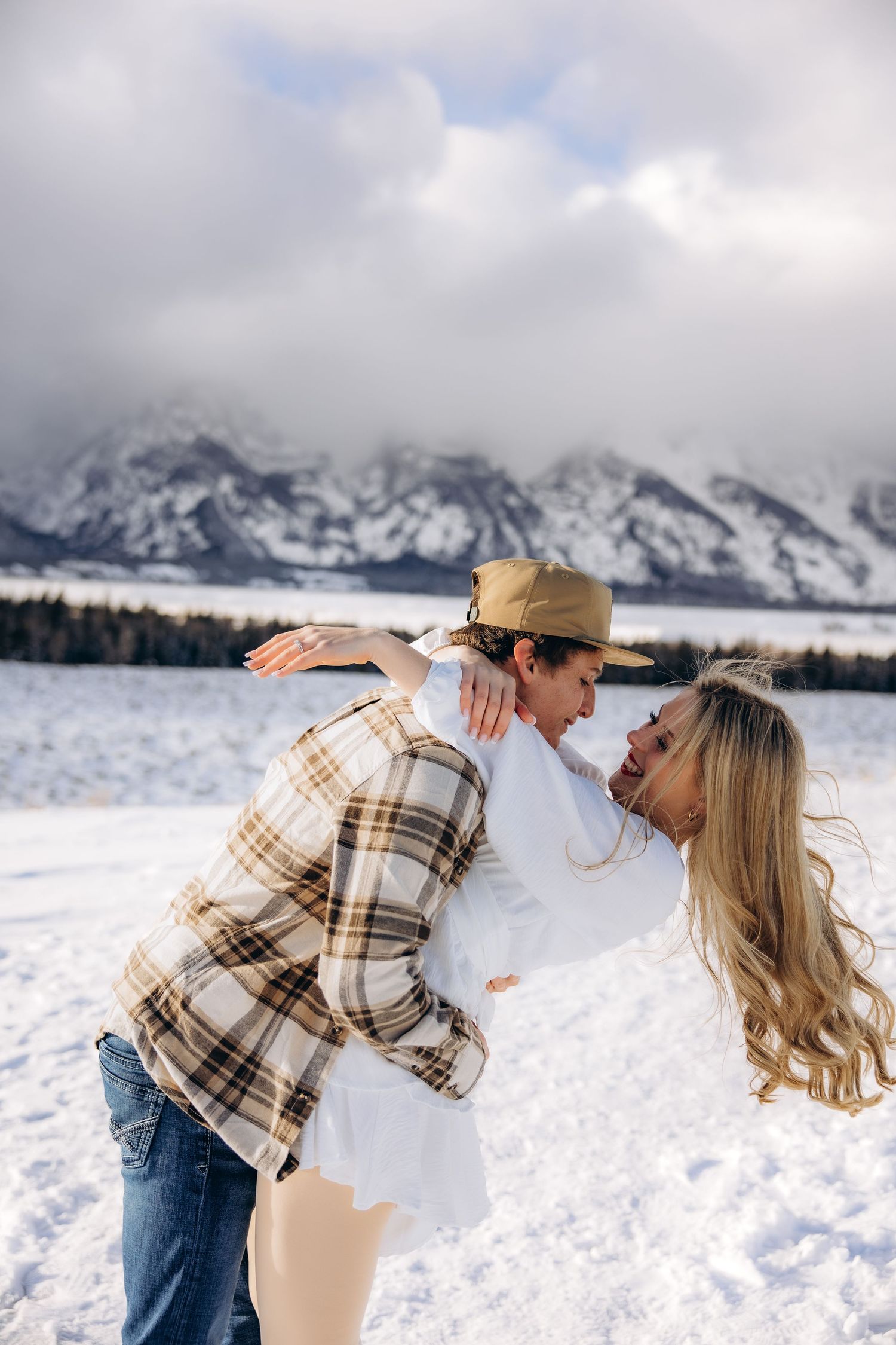 Romantic winter moment with snowy Teton mountains in background as couple embraces in plaid and denim.