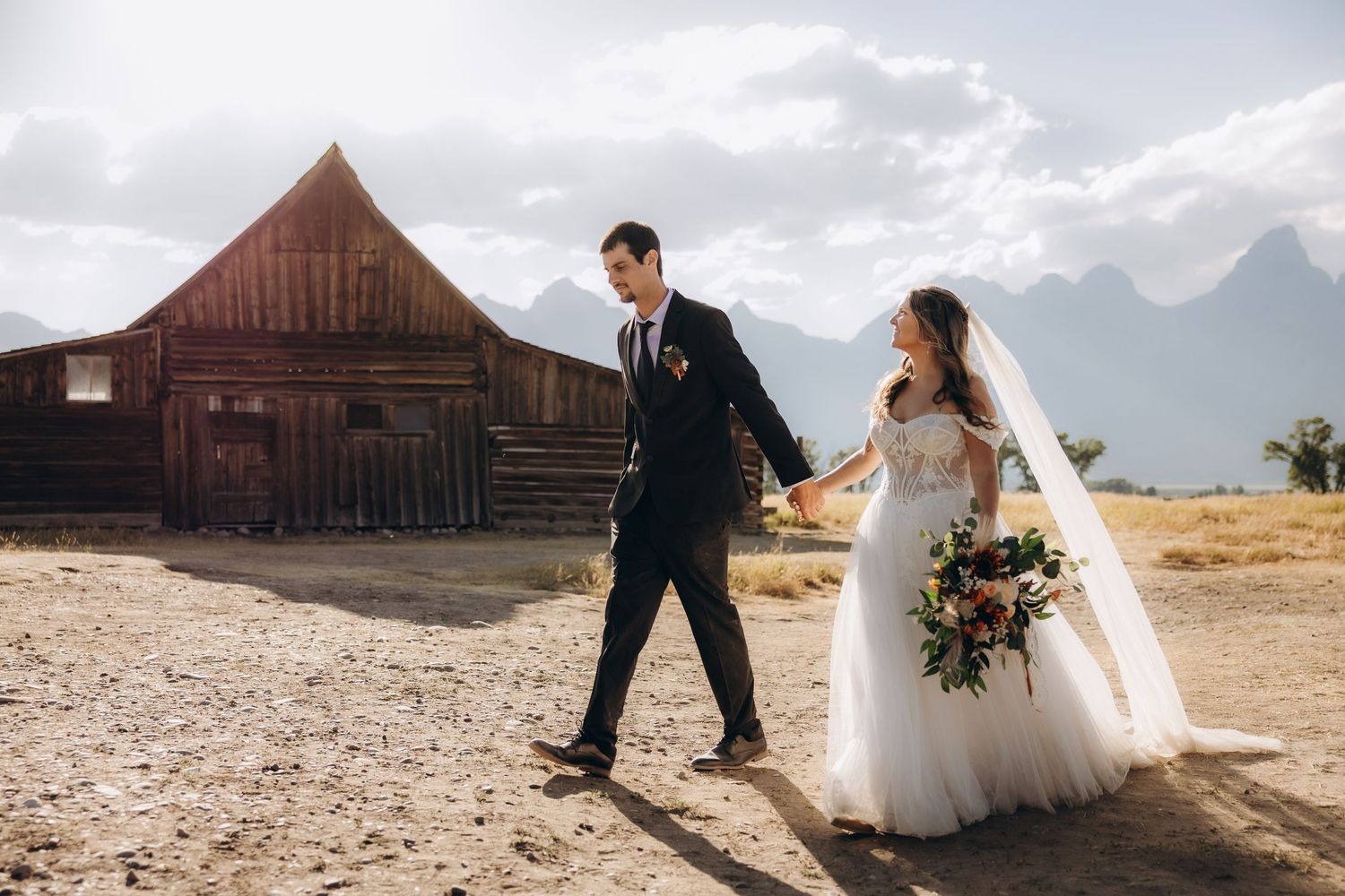 A couple in wedding attire stands in front of rustic barns and mountains in Grand Teton National Park during sunset.