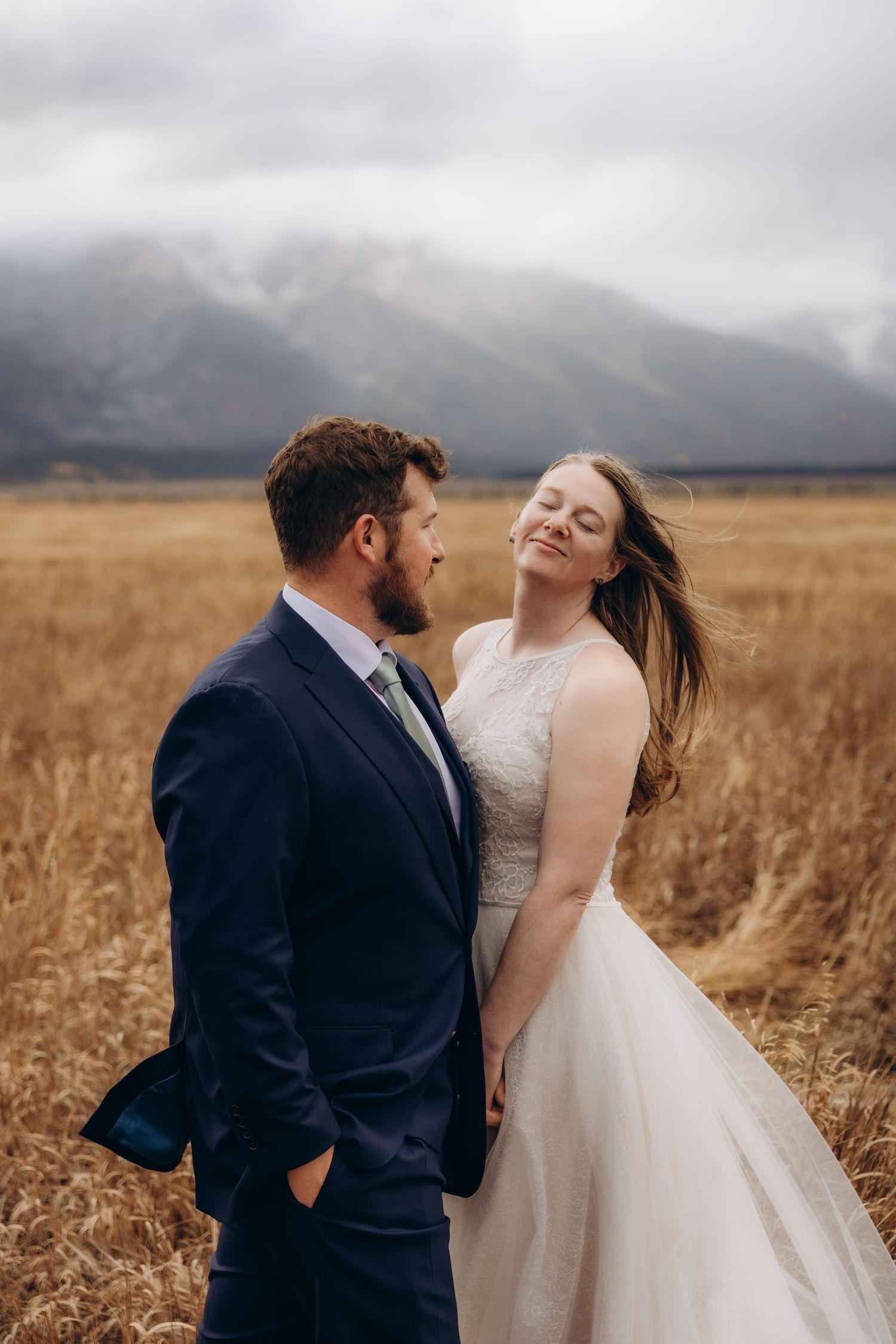 A couple in wedding attire shares a joyful moment together in a golden wheat field under moody storm clouds.