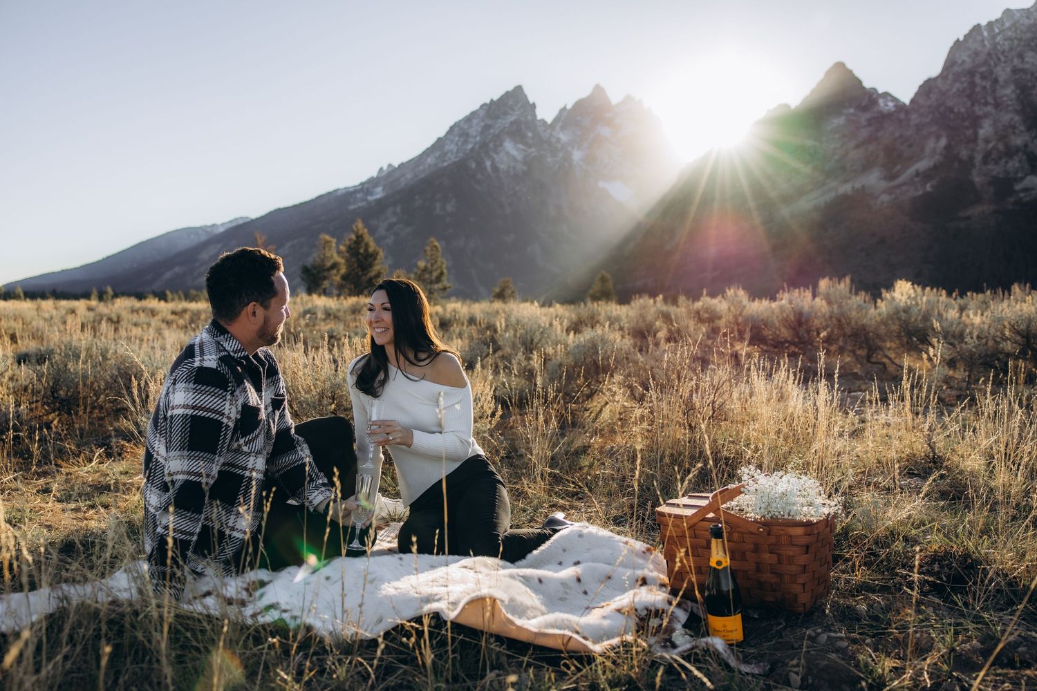 Two people sharing a romantic picnic on a blanket in a golden field with mountain peaks and sunburst in the background.