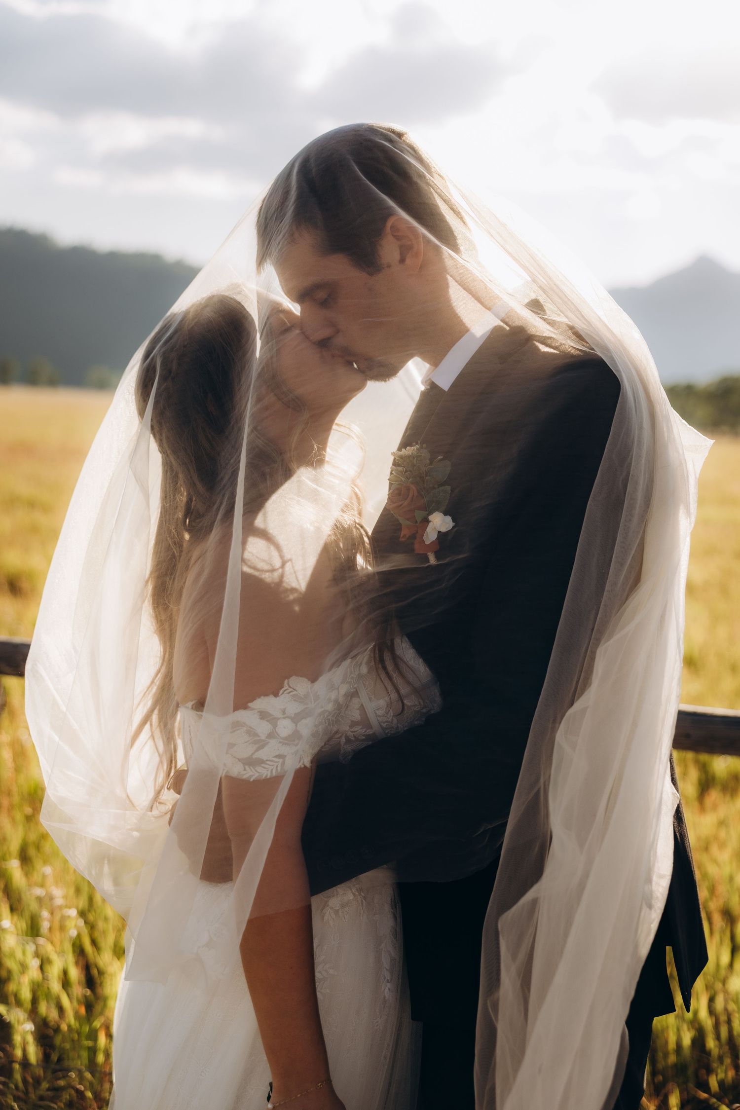 A romantic wedding couple embraces in a golden field at sunset as the bride's veil flows in the evening breeze.