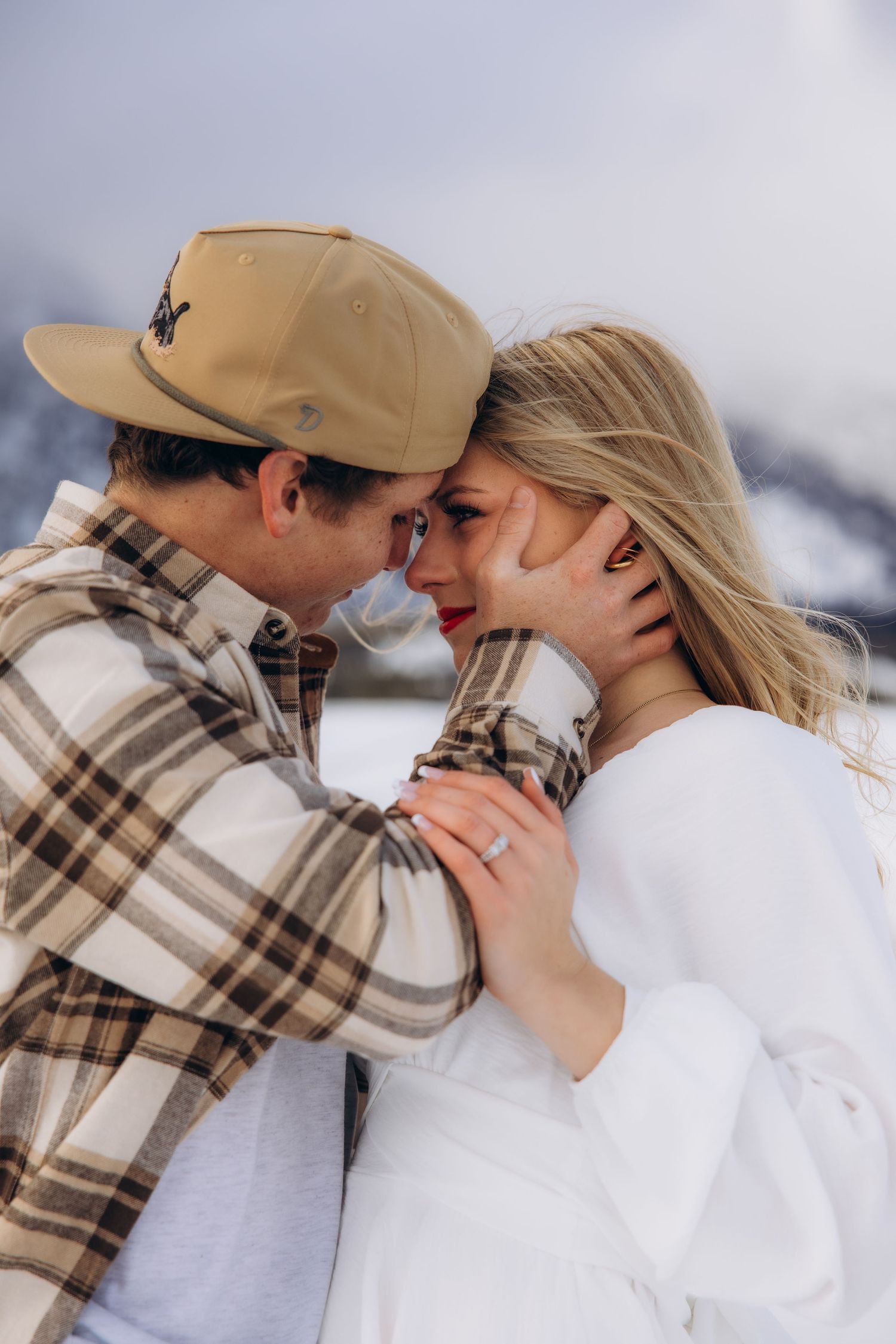 A couple shares an intimate moment in winter, wearing cozy sweaters against a snowy mountain backdrop.