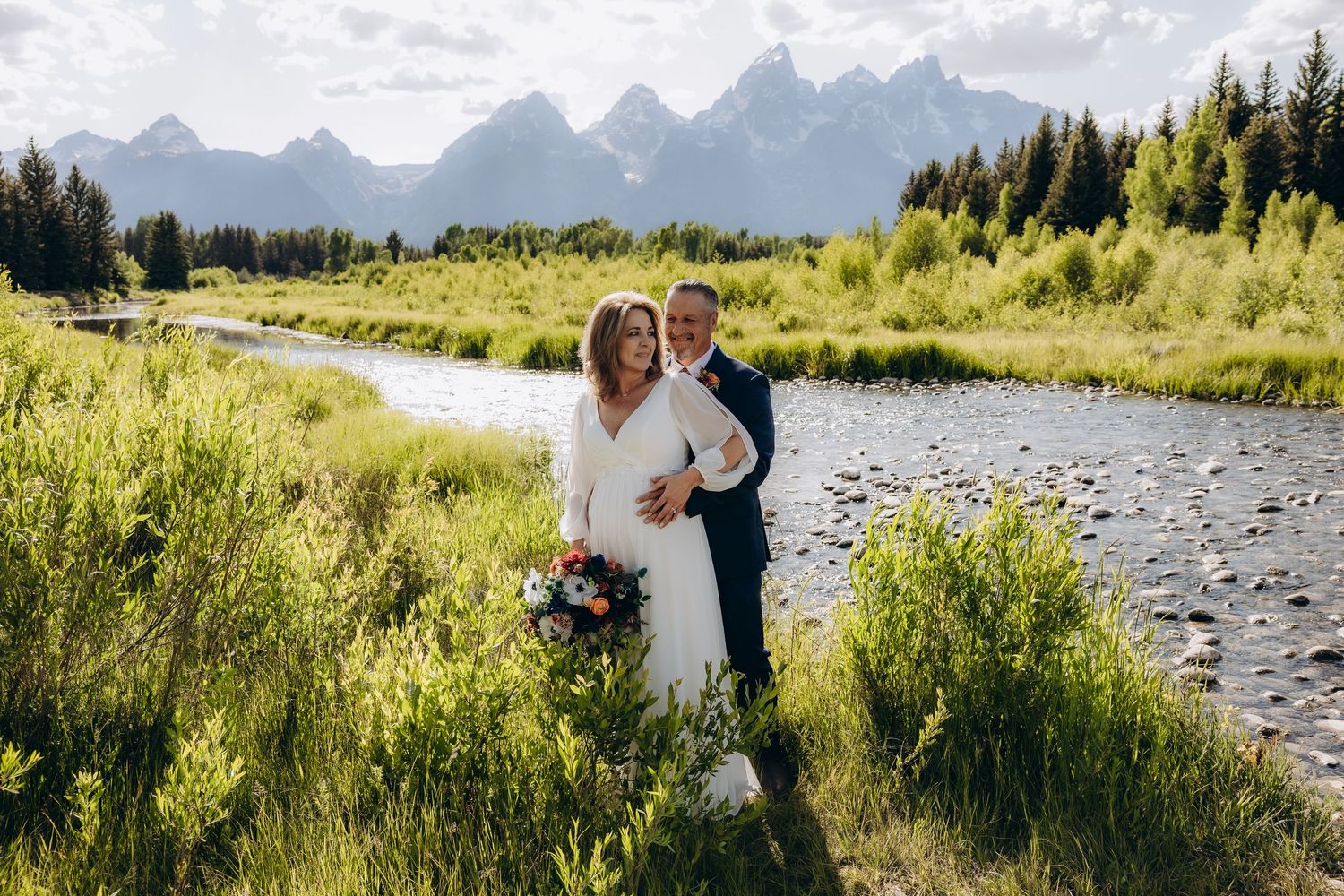 Couple shares romantic moments along a scenic mountain river surrounded by lush summer meadows and evergreen trees.