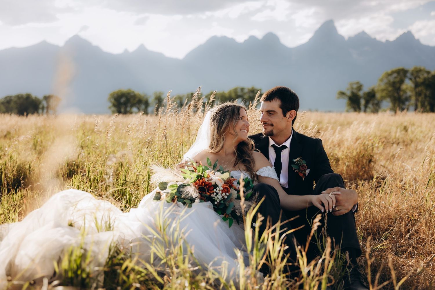 A romantic wedding photo in a golden field with mountain peaks in the background during sunset.
