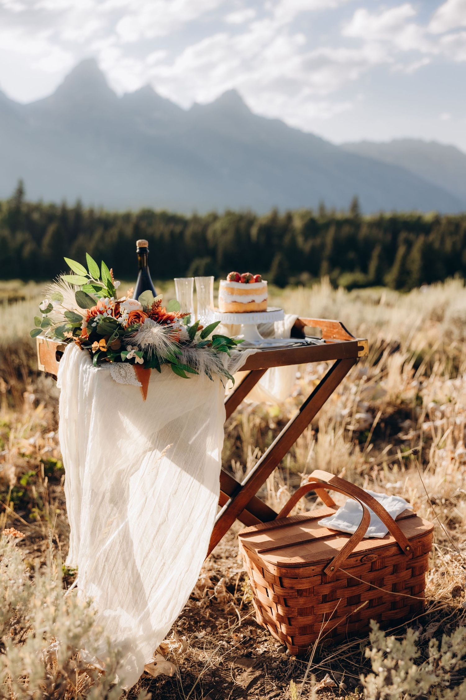 A decorative picnic basket adorned with floral arrangements sits in a golden field with majestic mountains in the background.