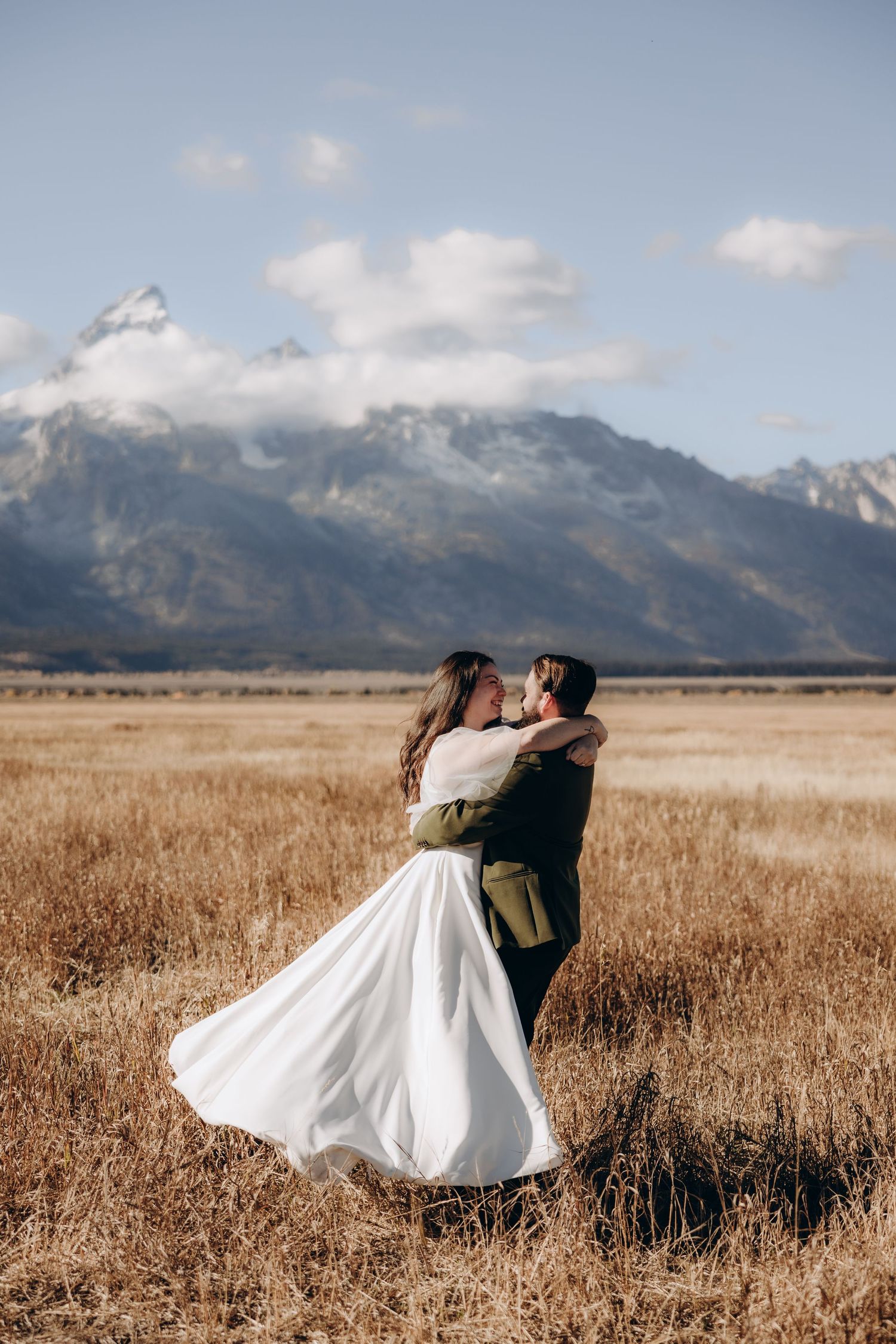 A couple in wedding attire embrace in a golden field with majestic snow-capped mountains in the background.