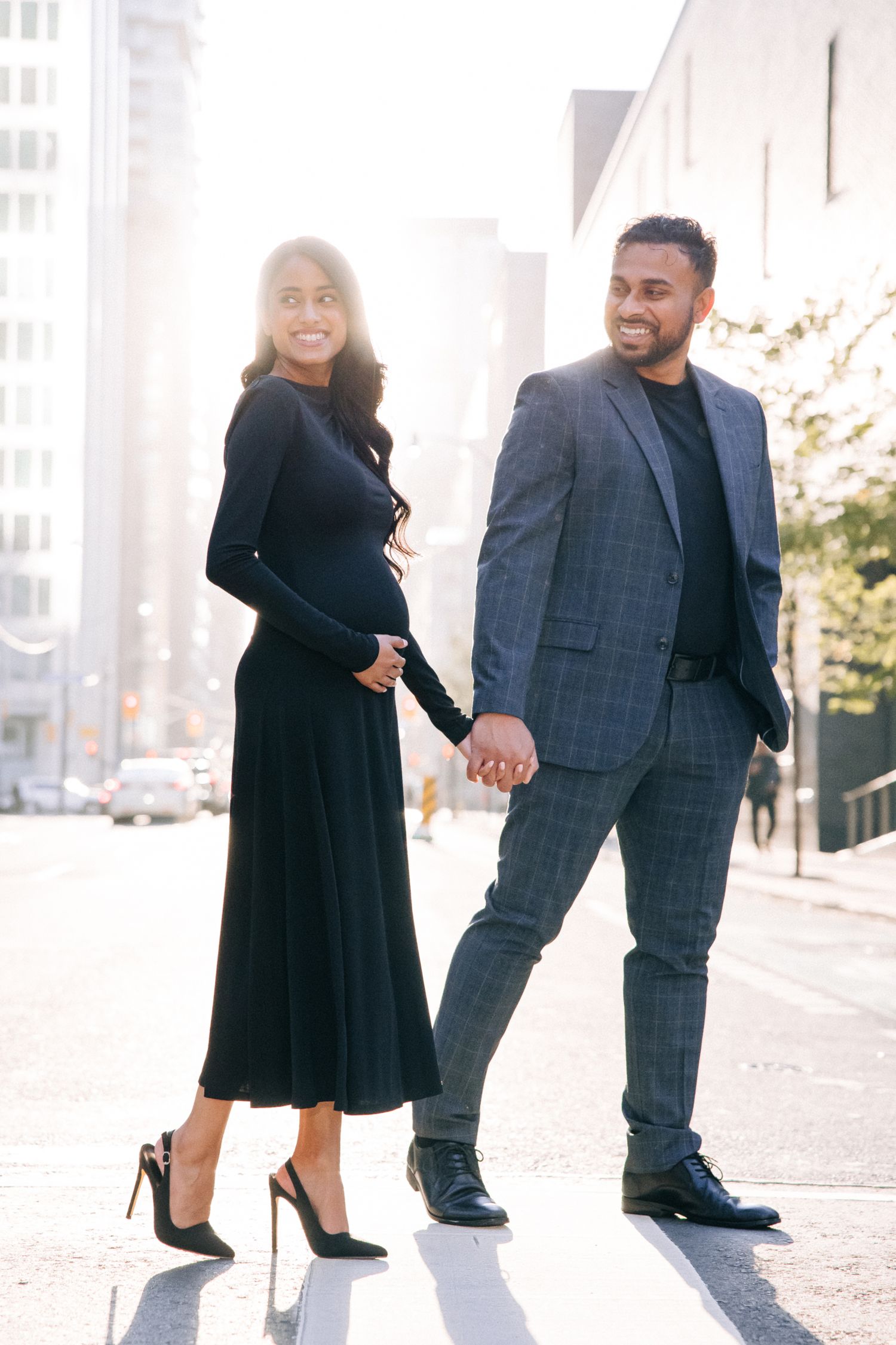 A couple holds hands while posing outdoors, with her wearing a black dress and him in a checkered suit.