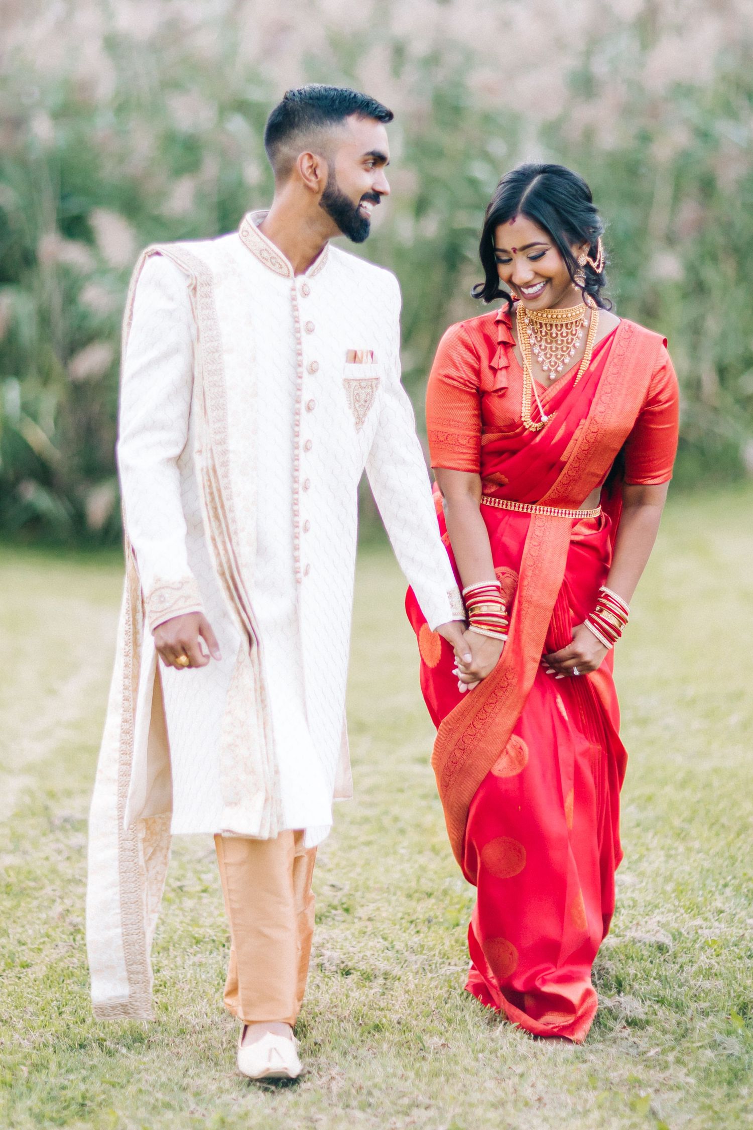 A couple in traditional South Asian wedding attire walking together and smiling in an outdoor garden setting.