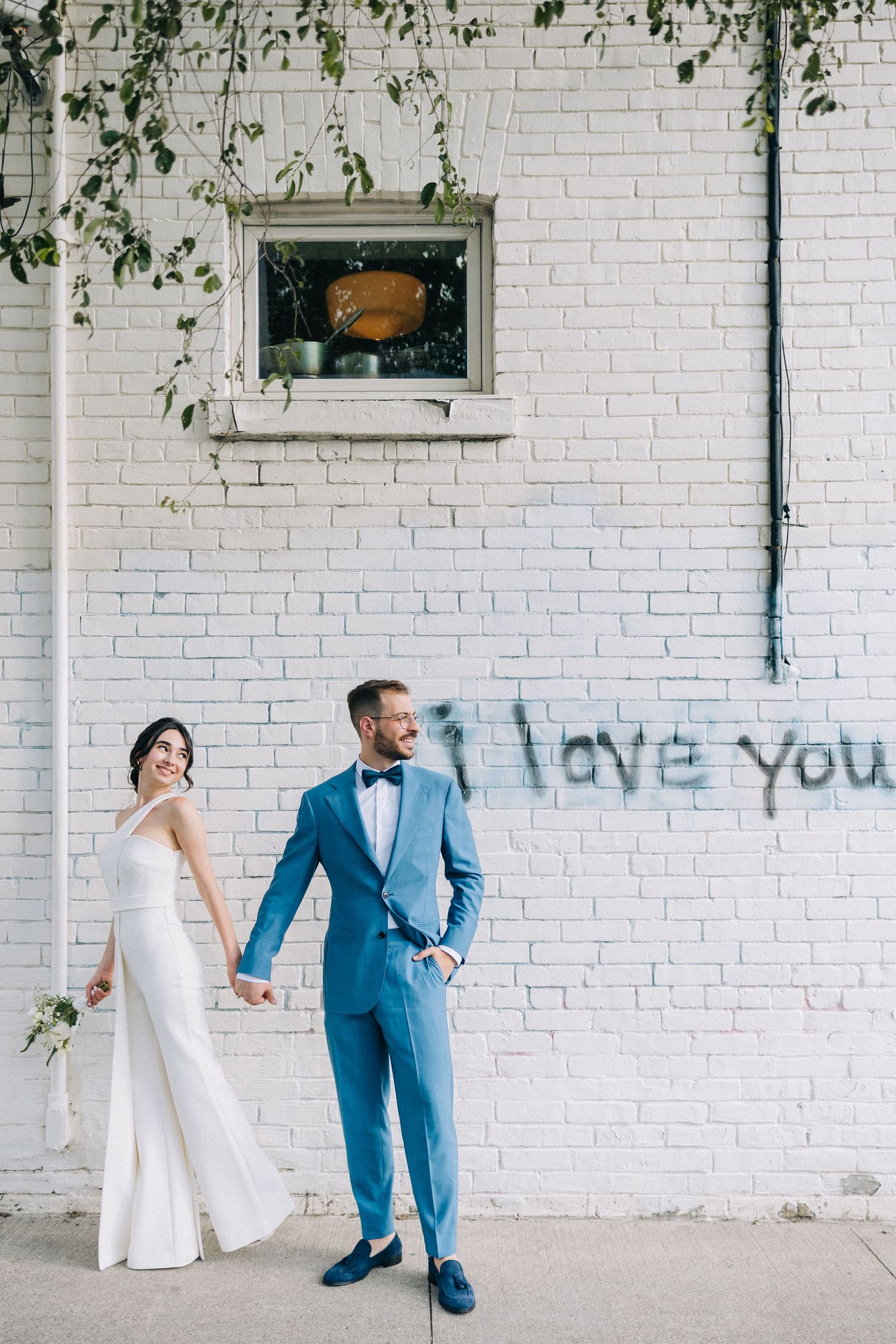 Couple in wedding attire poses against white brick wall with 