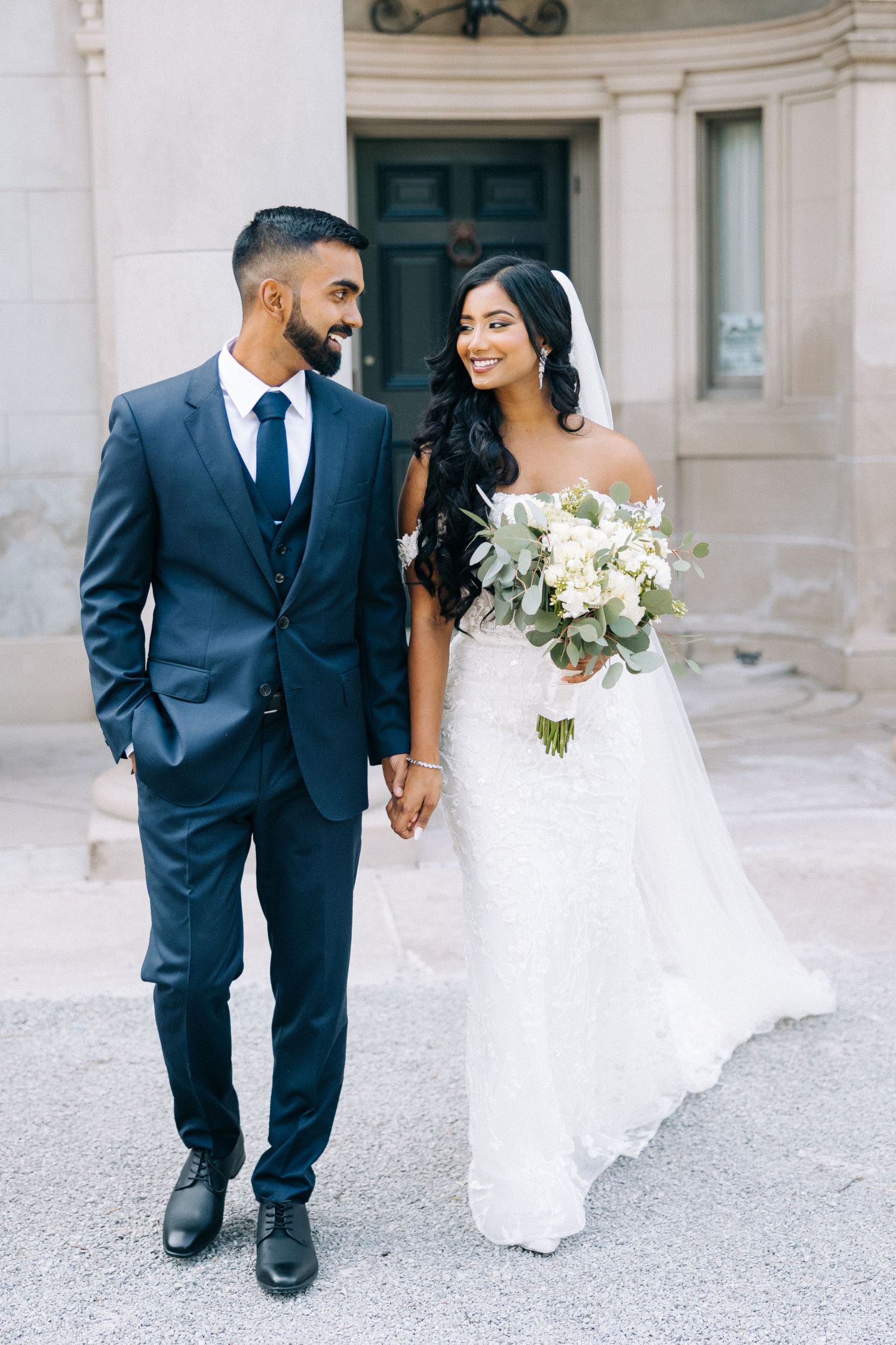 A couple walks hand in hand on their wedding day, with the bride in white and groom in navy blue suit.