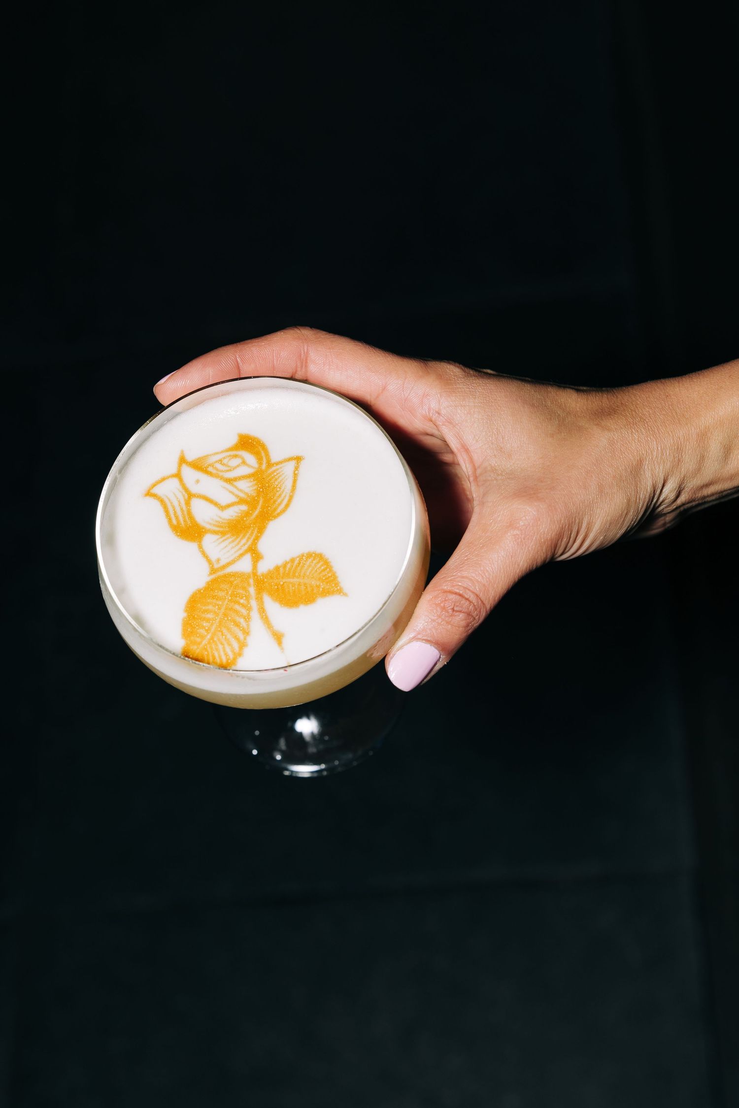 Cocktail with rose latte art design held against black background.