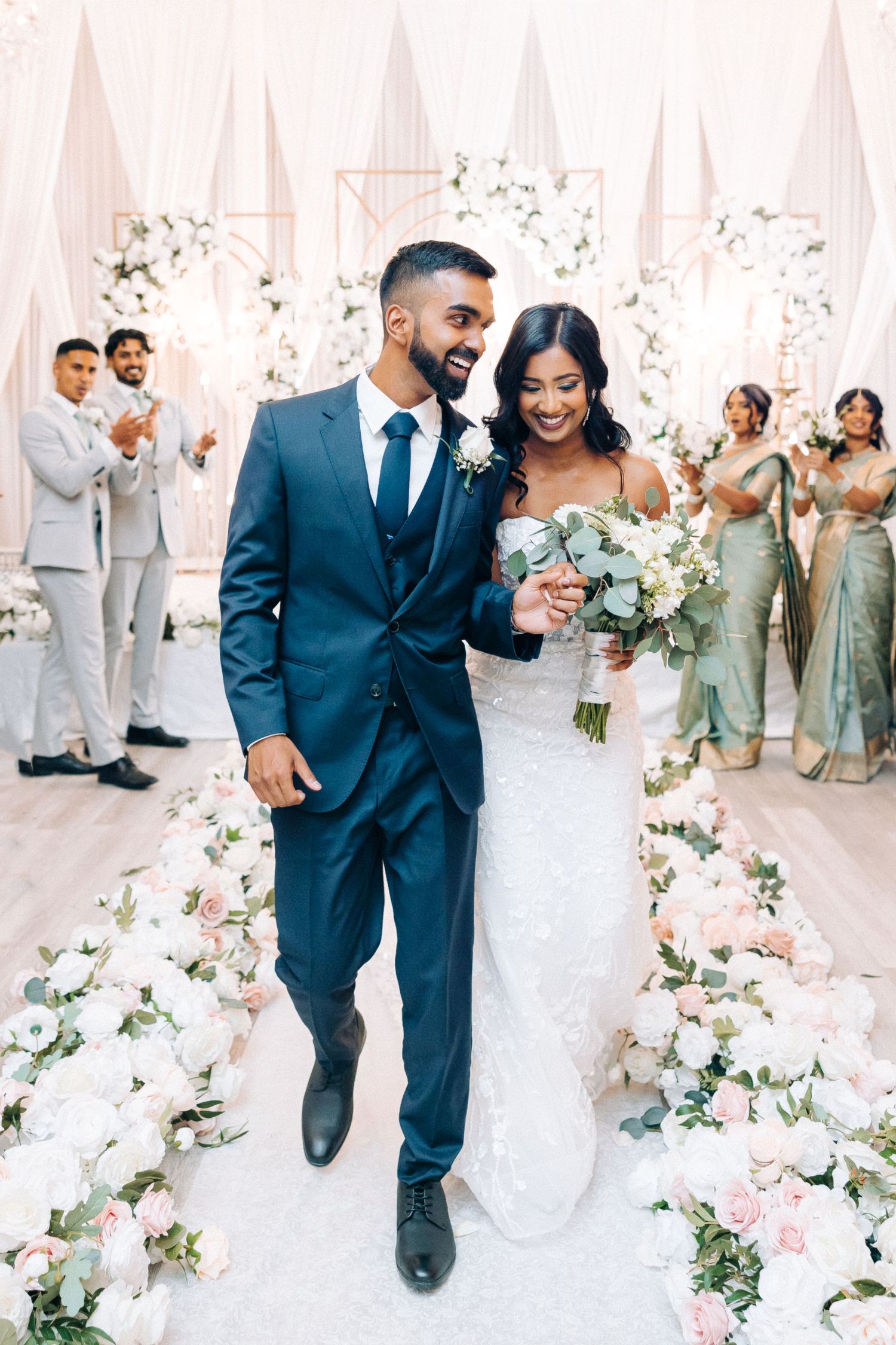 A joyful couple walks down a petal-lined aisle at their elegant wedding with light pink and white floral decorations.