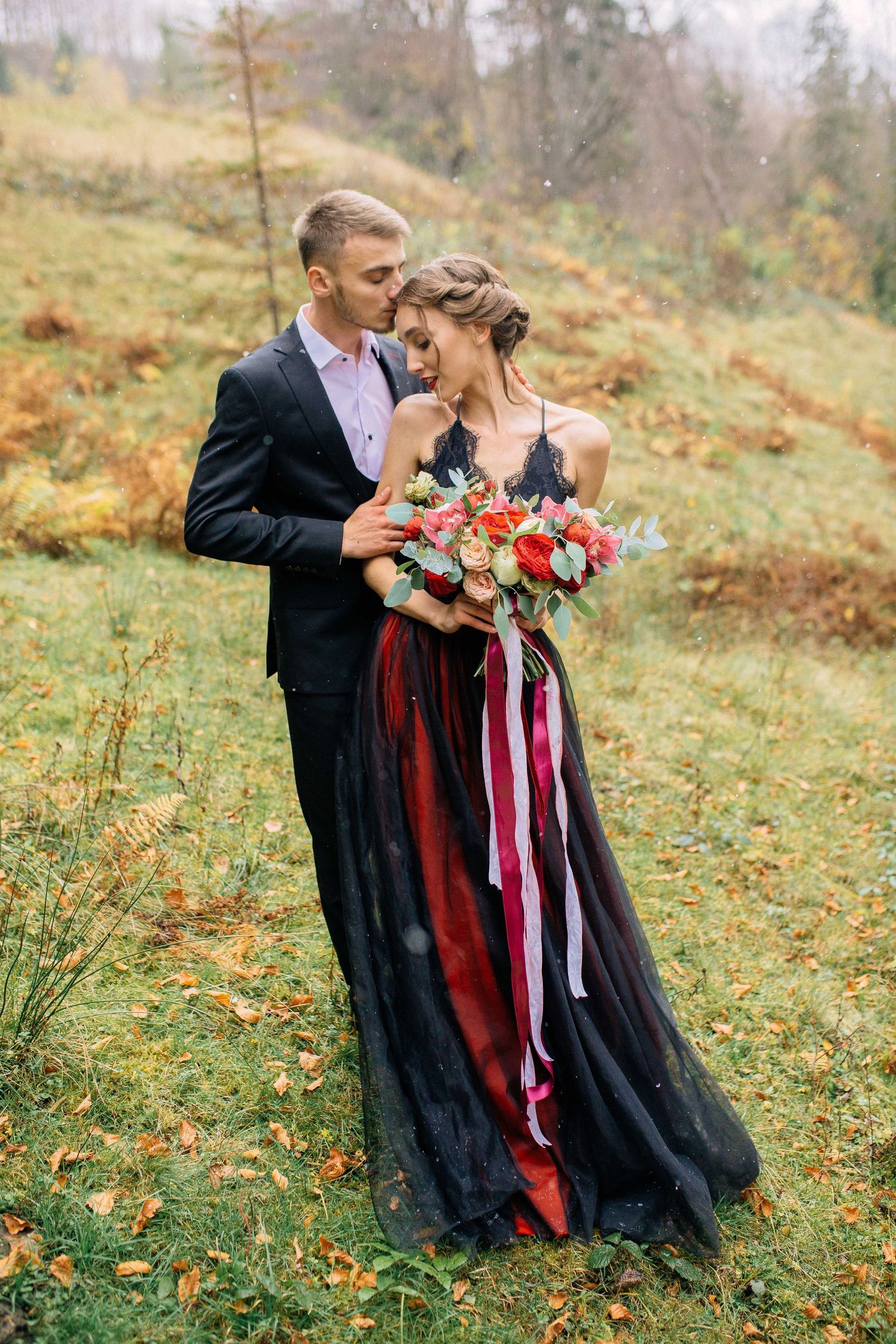 Romantic wedding couple in formal attire embraces on misty autumn hillside with burgundy and coral floral bouquet.