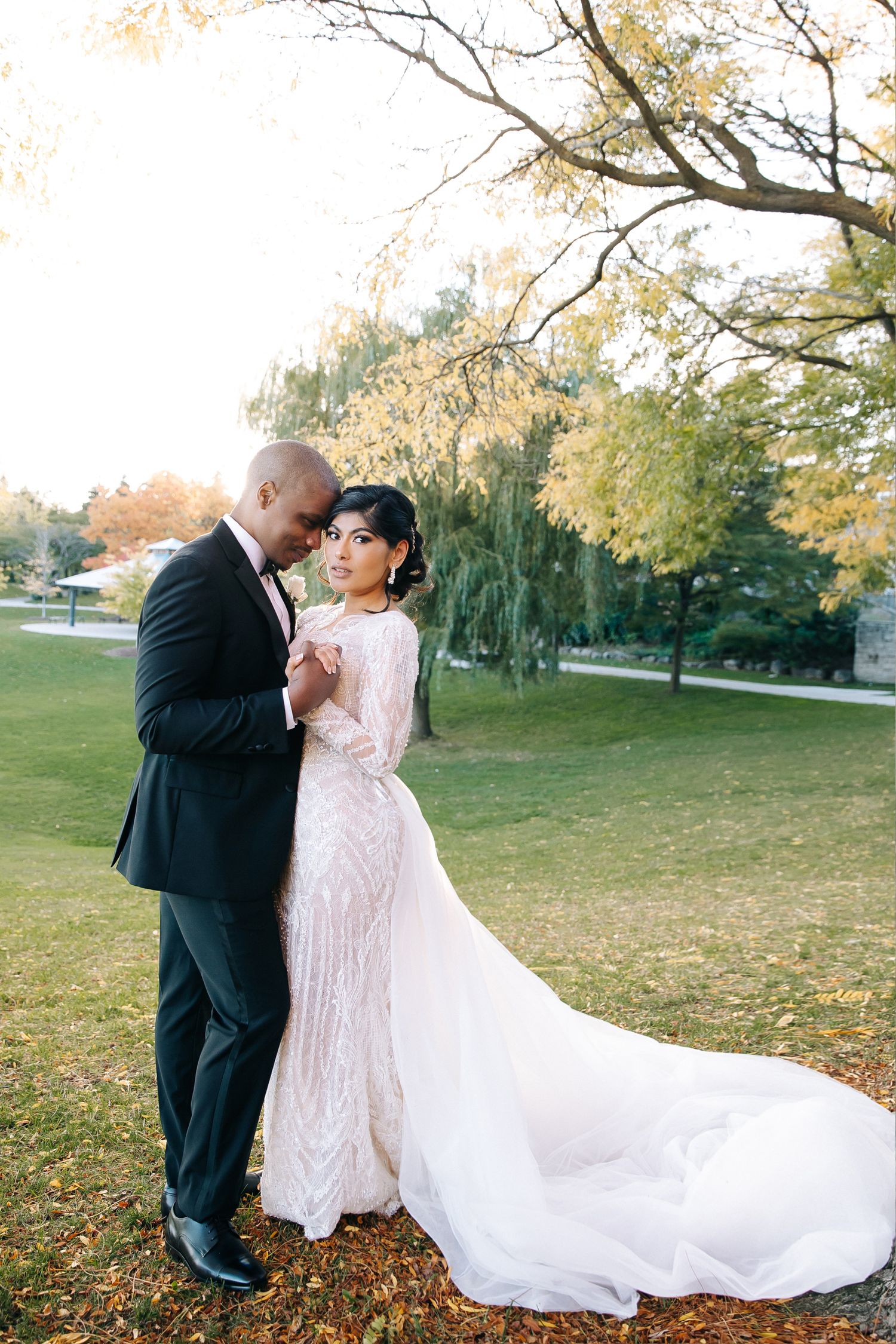 A romantic wedding couple embraces outdoors during autumn with a flowing white gown and fall foliage in the background.