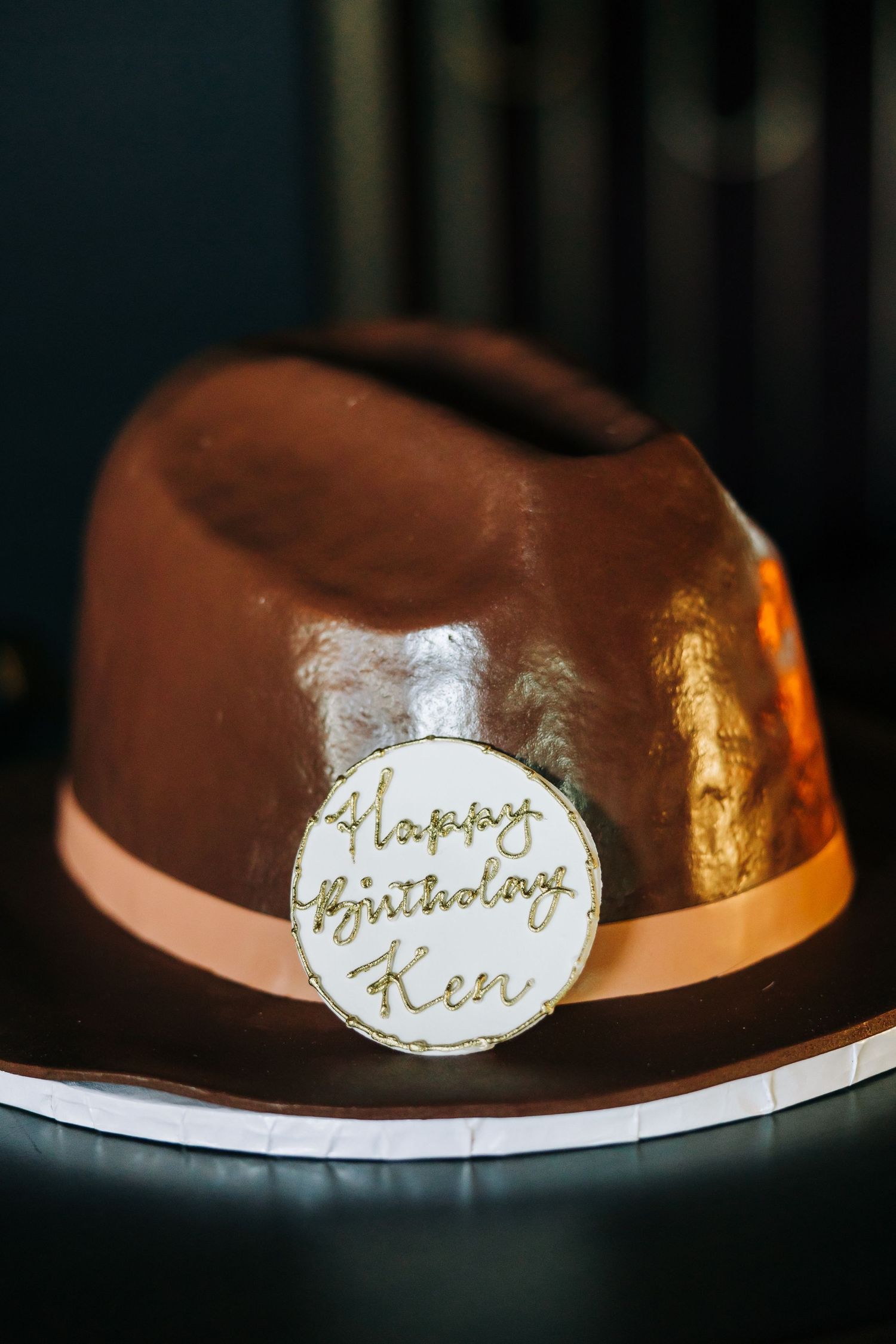A brown chocolate birthday cake with a round white fondant disc featuring gold 'Happy Birthday' text and gold accents.