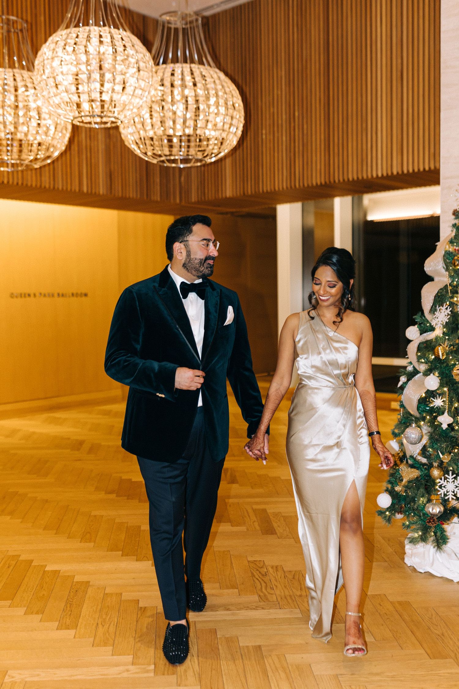An elegantly dressed couple walks hand in hand past a decorated Christmas tree in a luxurious venue.