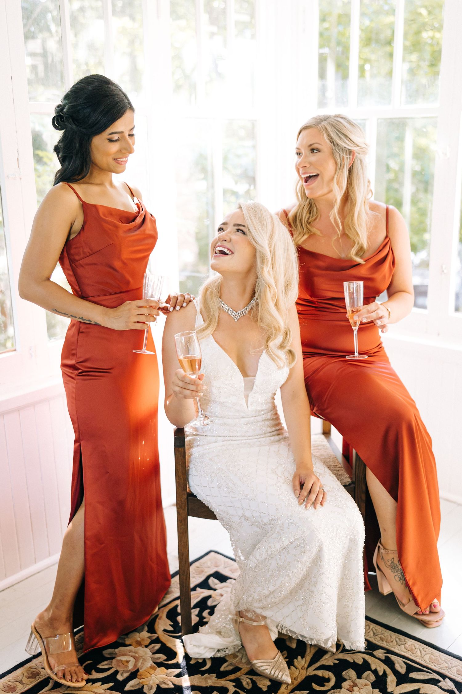 Three women share joyful laughter while wearing elegant dresses in rust orange and white at a formal event.