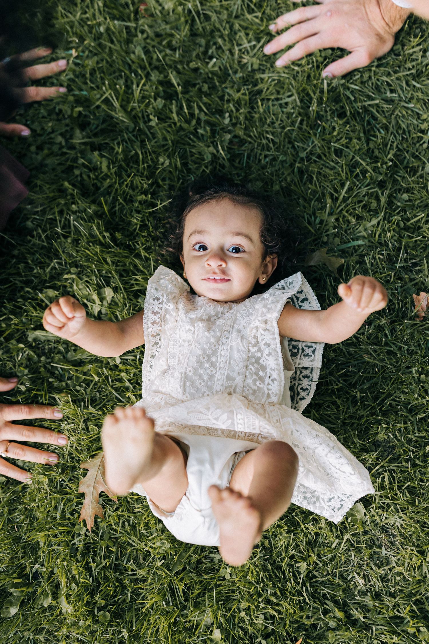 Baby in white lace dress lying on green grass with hands reaching towards them from outside the frame.