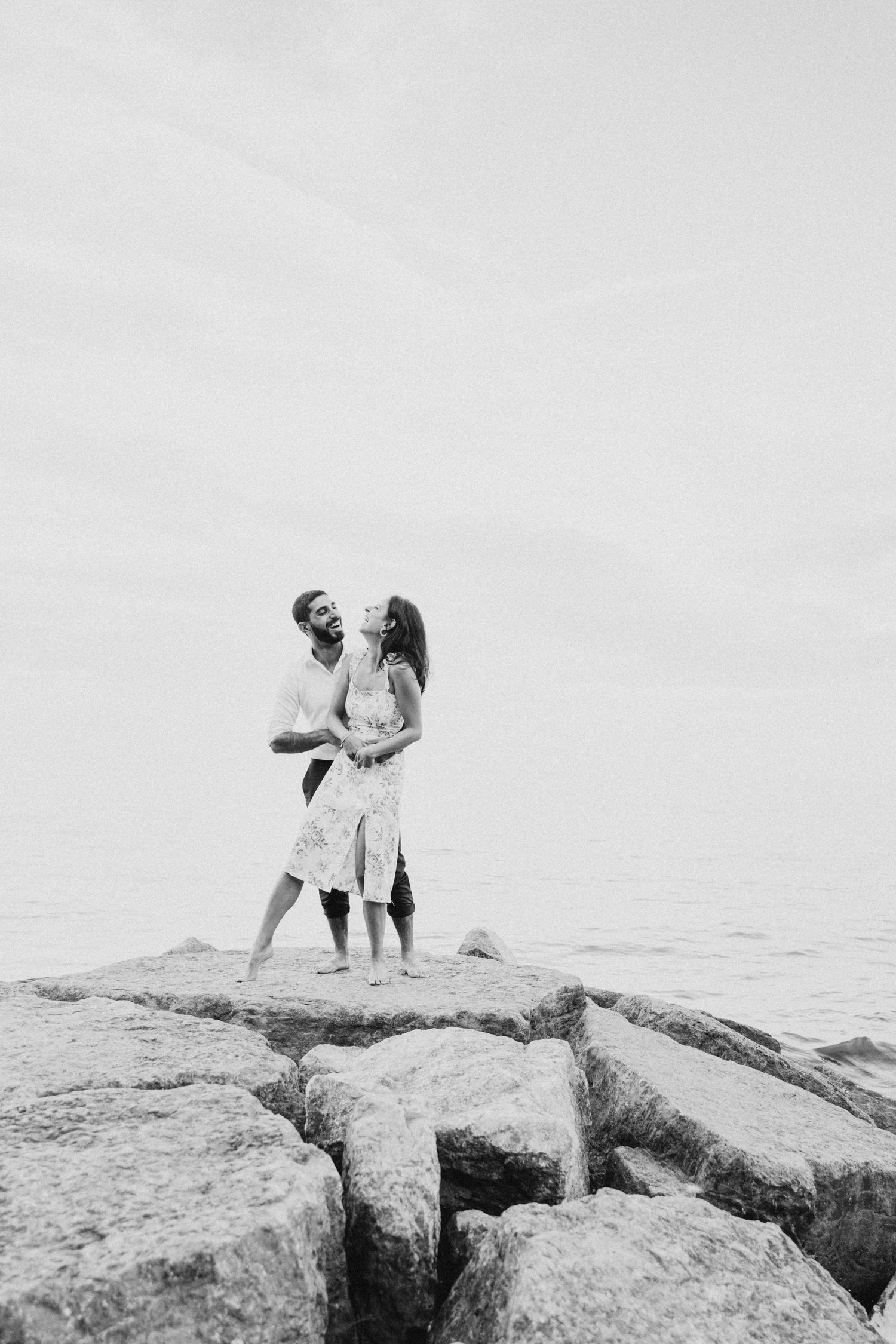 A romantic couple stands together on rocky coastal outcrop in black and white photography.