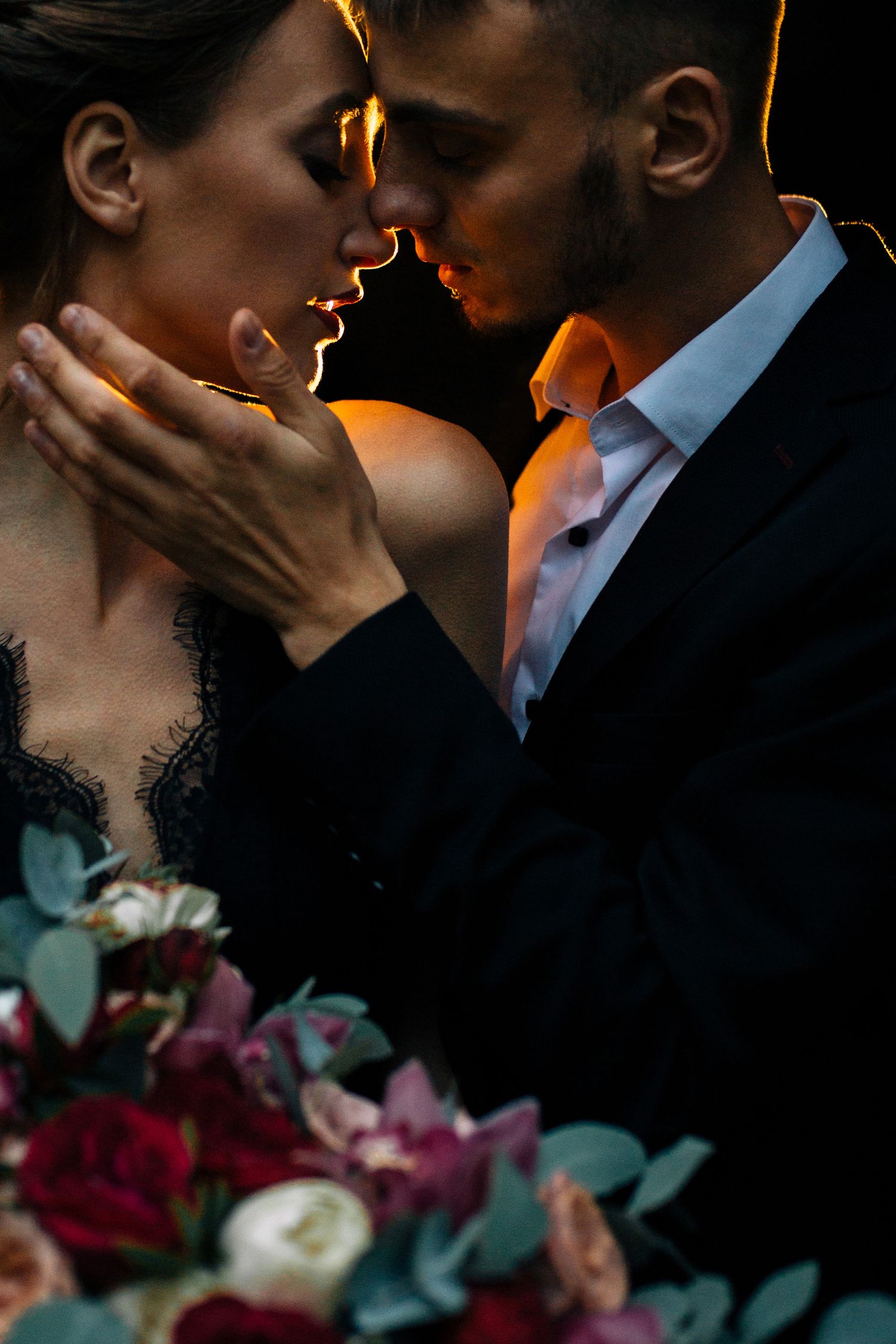 Intimate couple in romantic pose with dramatic lighting and vibrant floral bouquet in foreground.