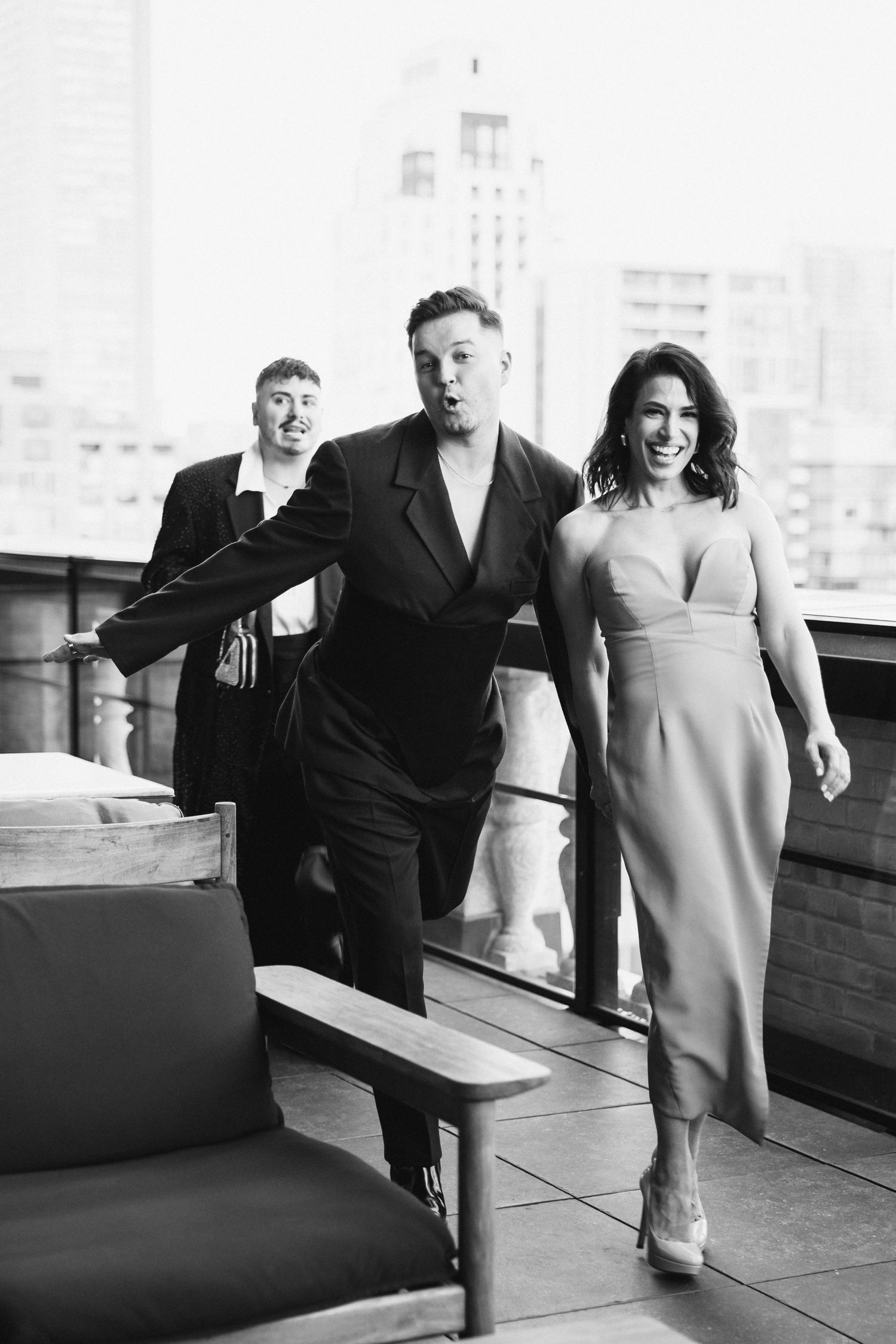 Black and white photo of people walking together on a rooftop terrace in formal attire with city buildings in background.