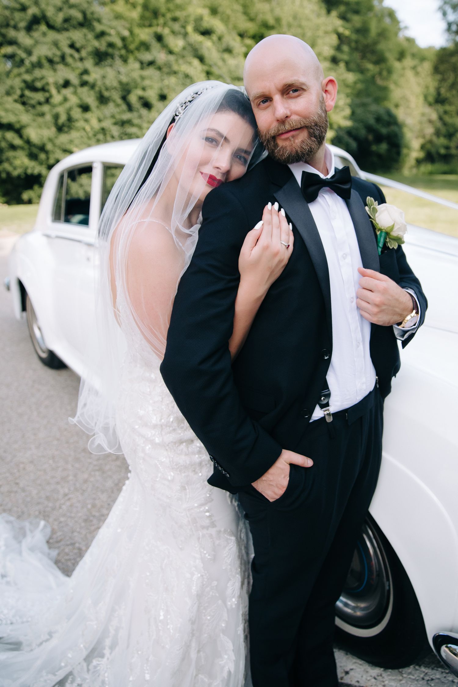 Happy newlyweds pose by vintage car after wedding ceremony in outdoor setting with lush greenery in background.