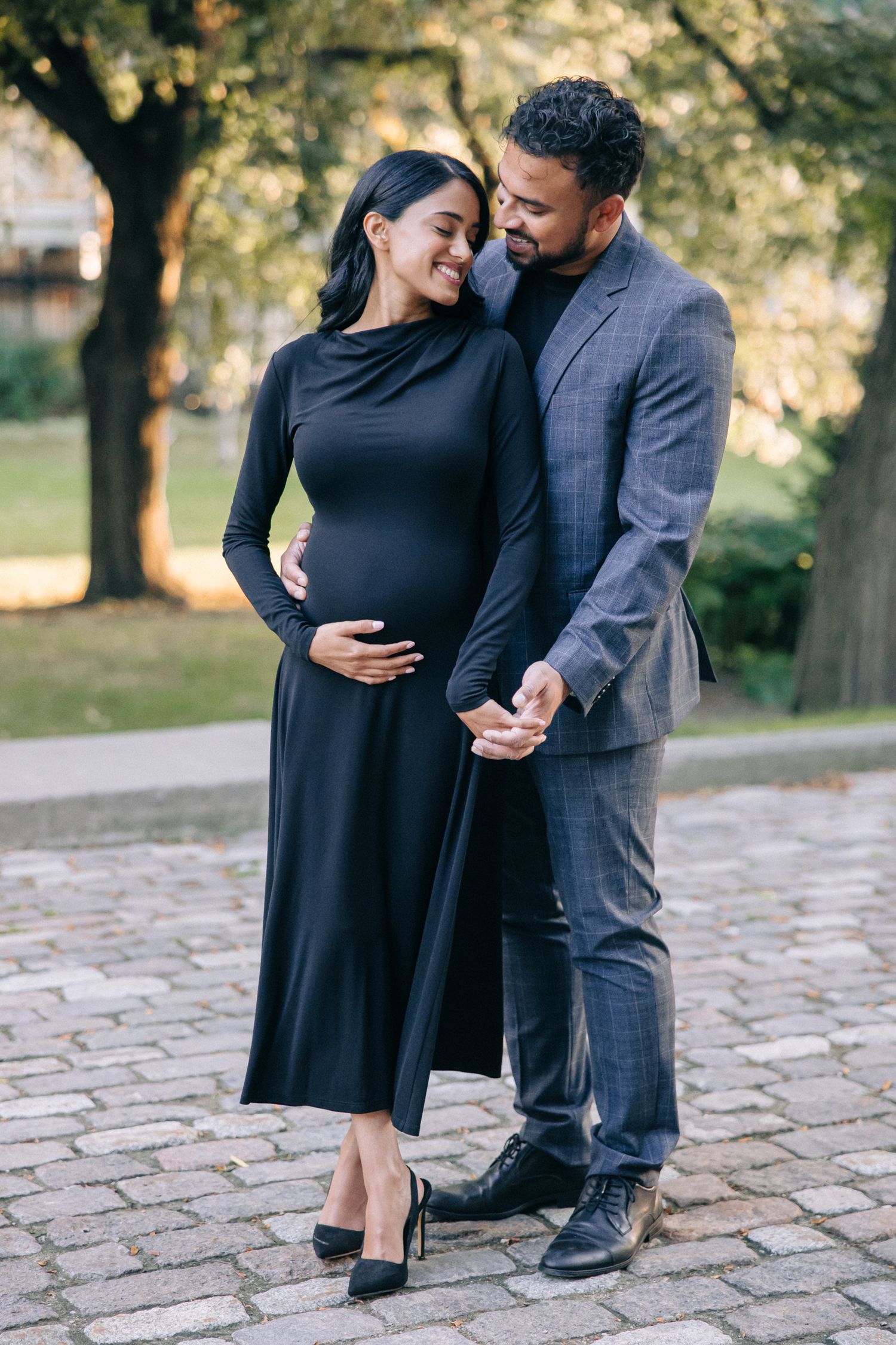 A couple shares an intimate moment outdoors, with her in a black dress touching her pregnant belly.