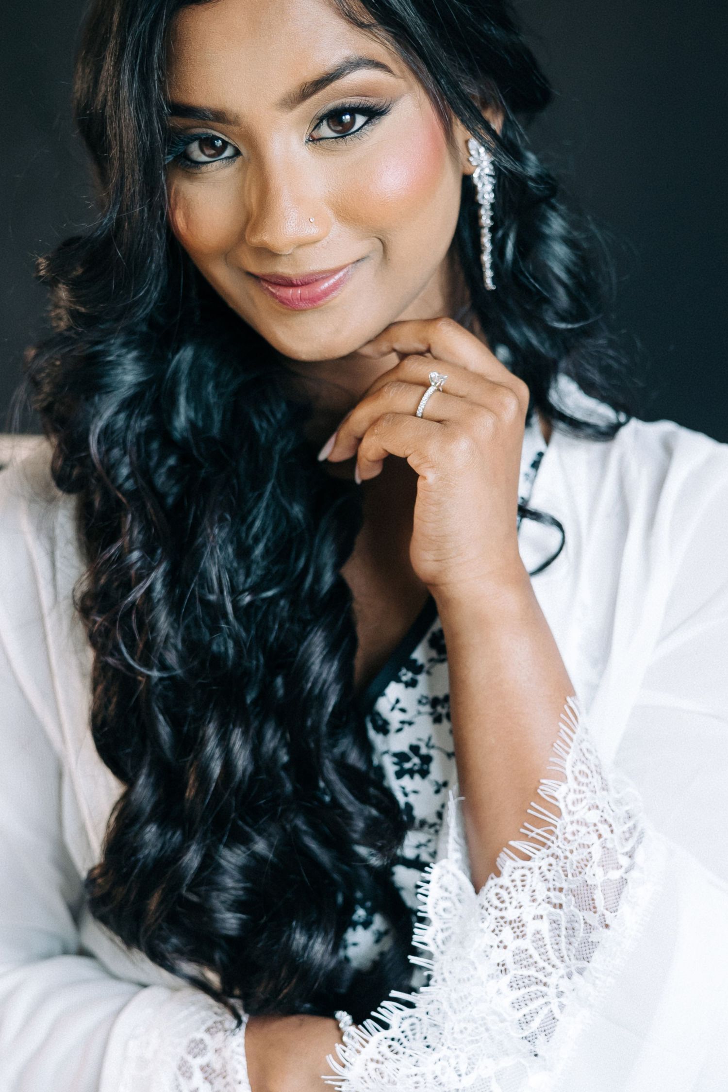 Long wavy black hair and sparkling earrings complement an elegant white lace robe in this glamorous beauty portrait.