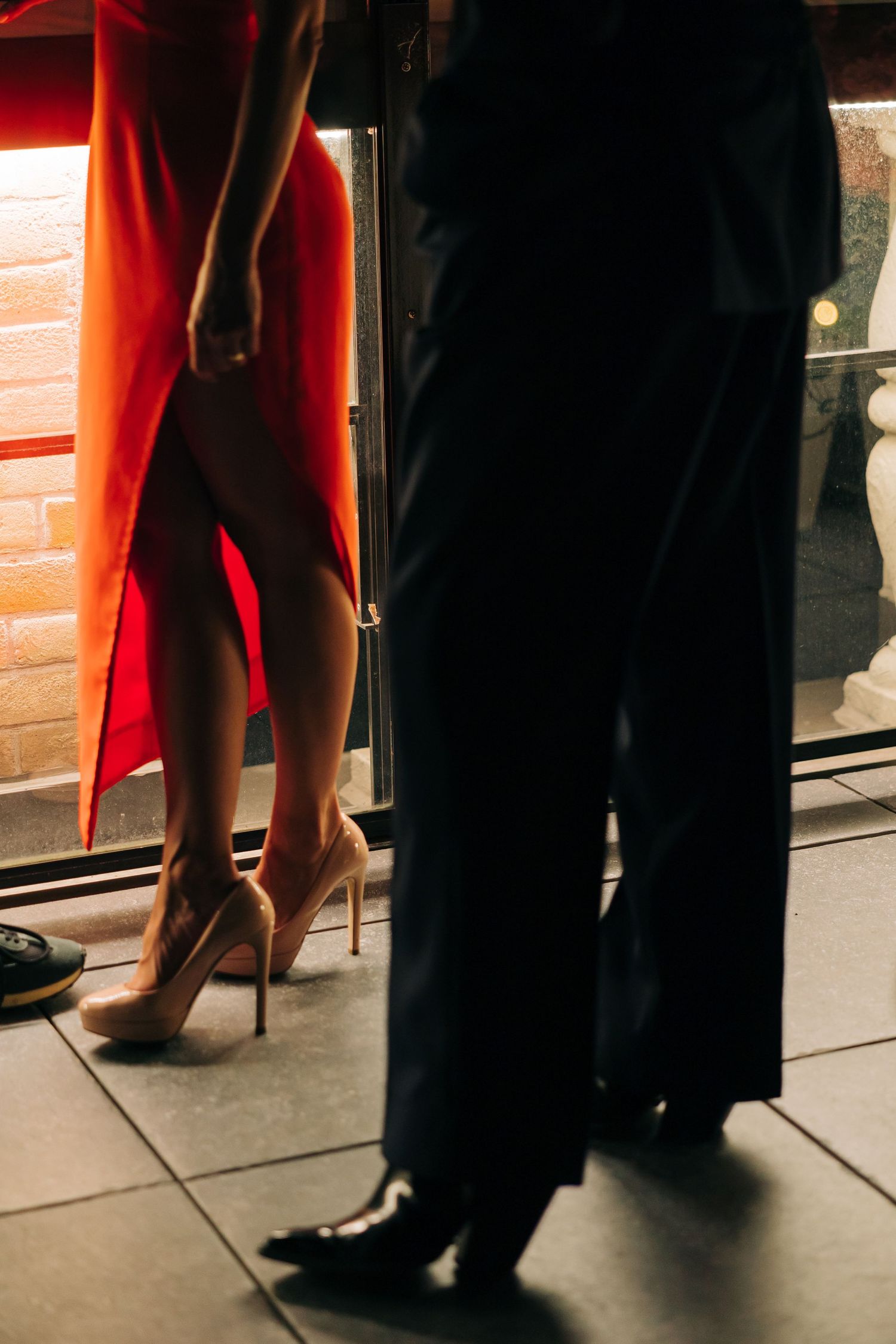 A close-up of two contrasting outfits, with legs wearing red pants and nude heels next to dark formal trousers and dress shoes.