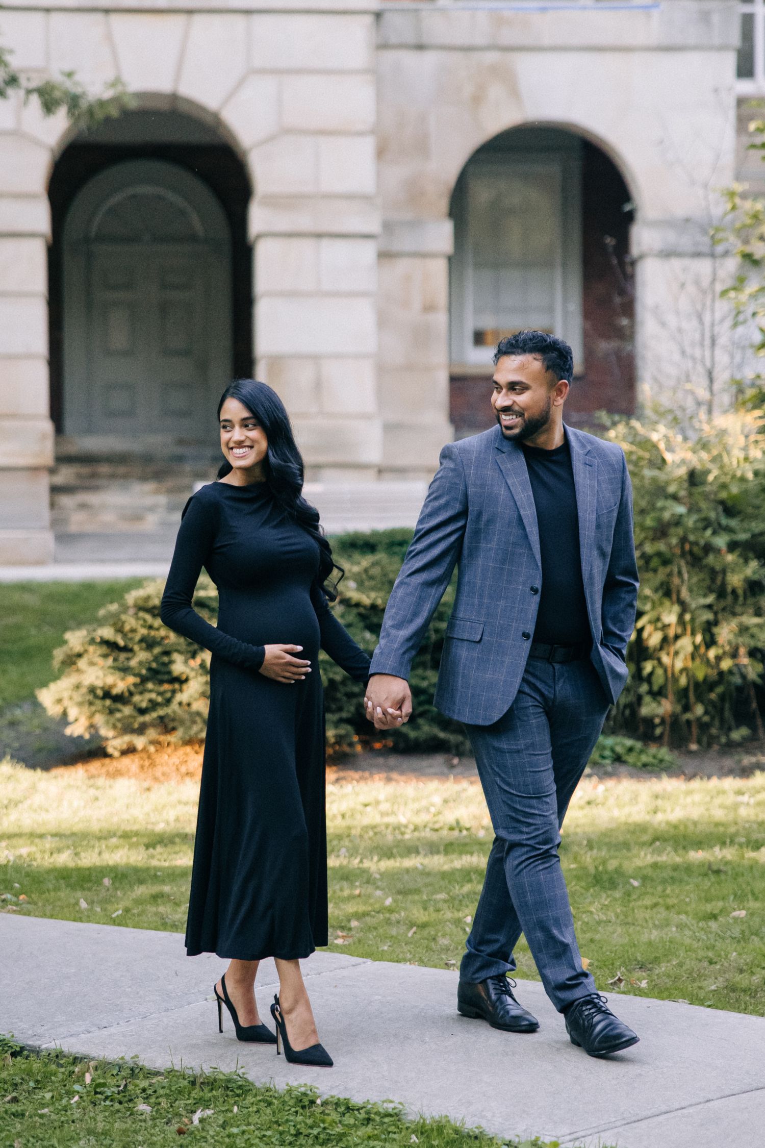 A couple walks hand in hand on a sidewalk, with the woman in a long black dress and man wearing a blue suit.