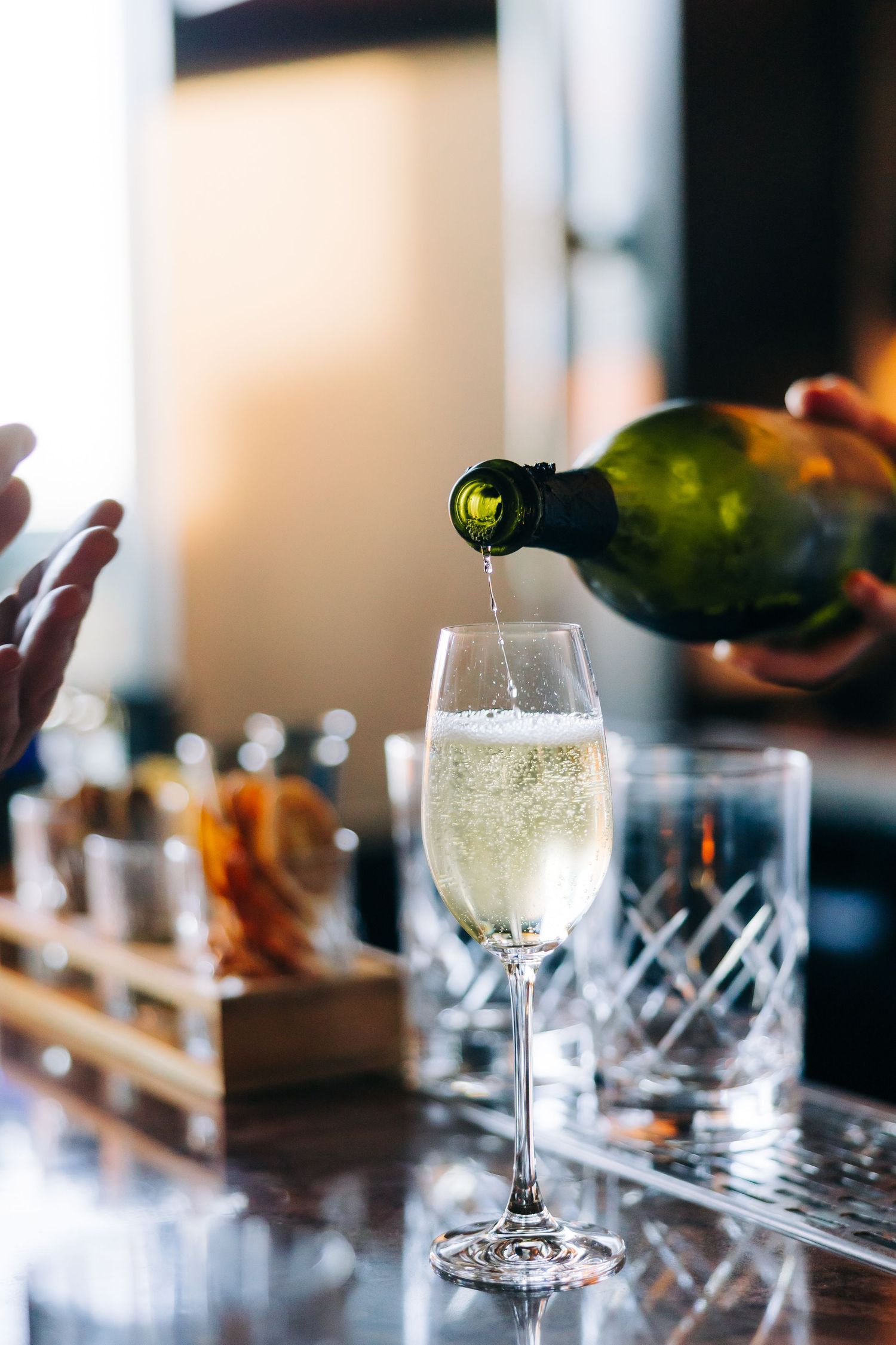 Champagne being poured into an elegant crystal glass at a bar with glasses and serving boards in the background.