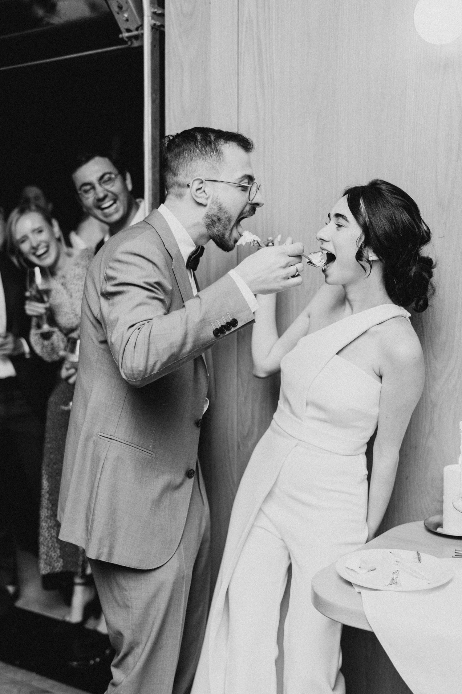 A newlywed couple playfully feed each other wedding cake while guests laugh in the background at a reception celebration.